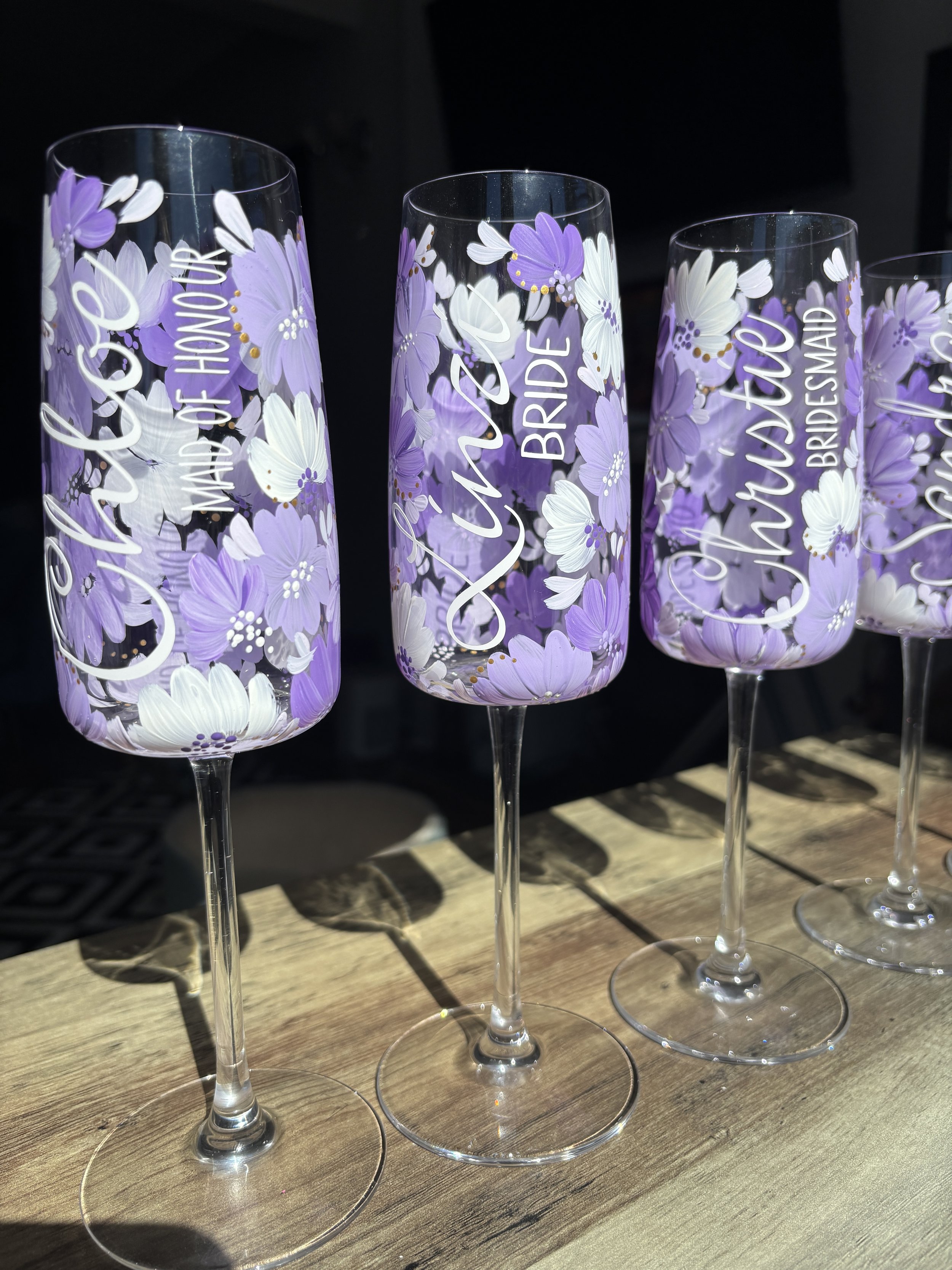 Four tall champagne flutes decorated with purple and white flowers, with names and wedding titles on them like "Bridesmaid", "Mother of the Groom", and "Maid of Honour".