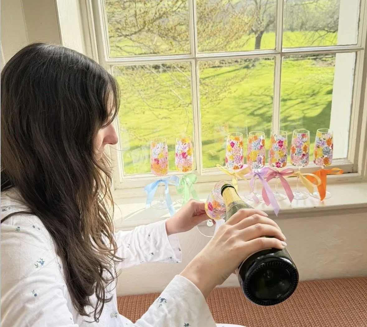 A bride with long dark hair pouring champagne into a painted flute glass with a floral design, while sitting near a window with a view of green trees and grass.