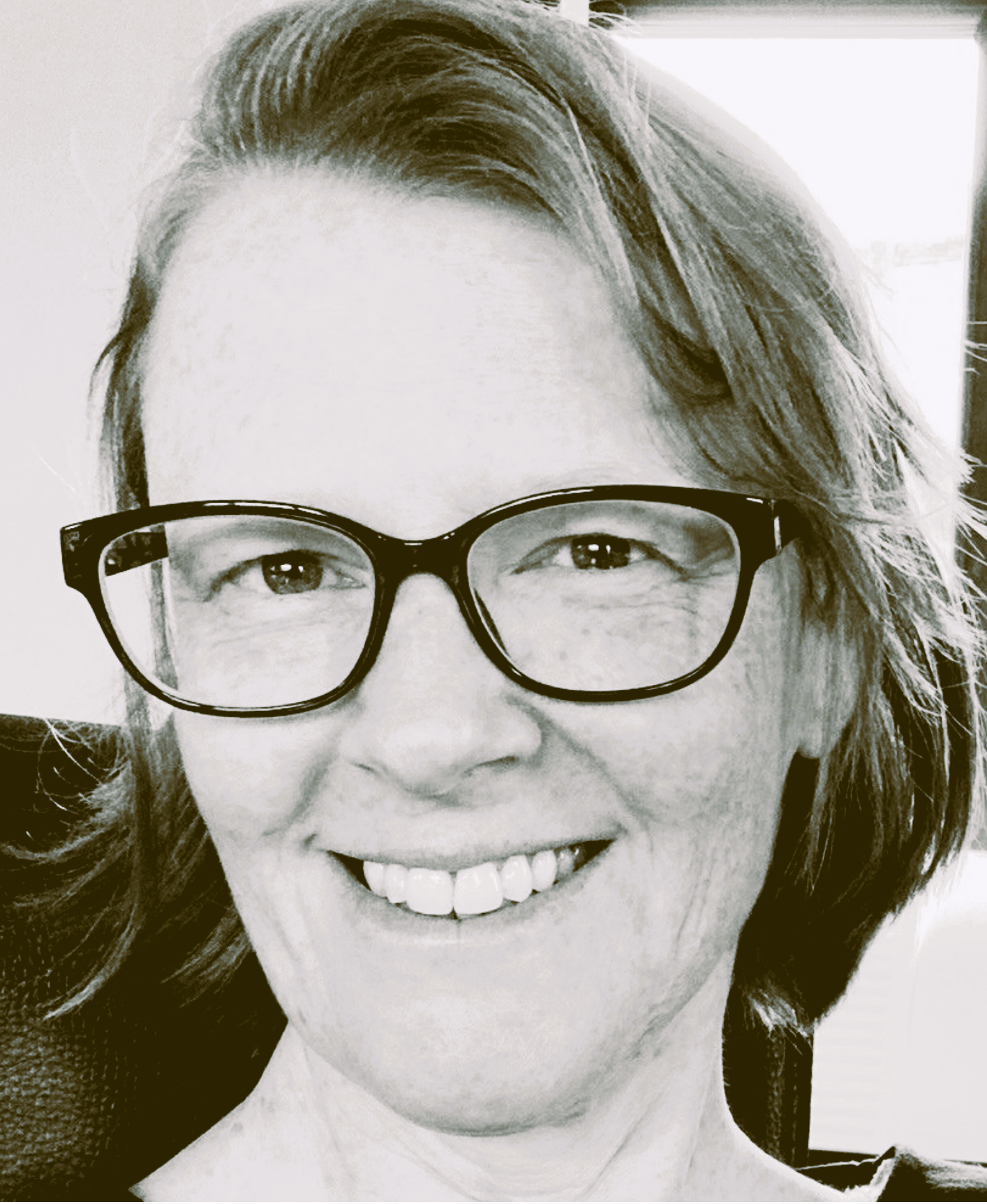 Close-up black and white photograph of a woman with glasses, smiling, showing her teeth, with shoulder-length hair.