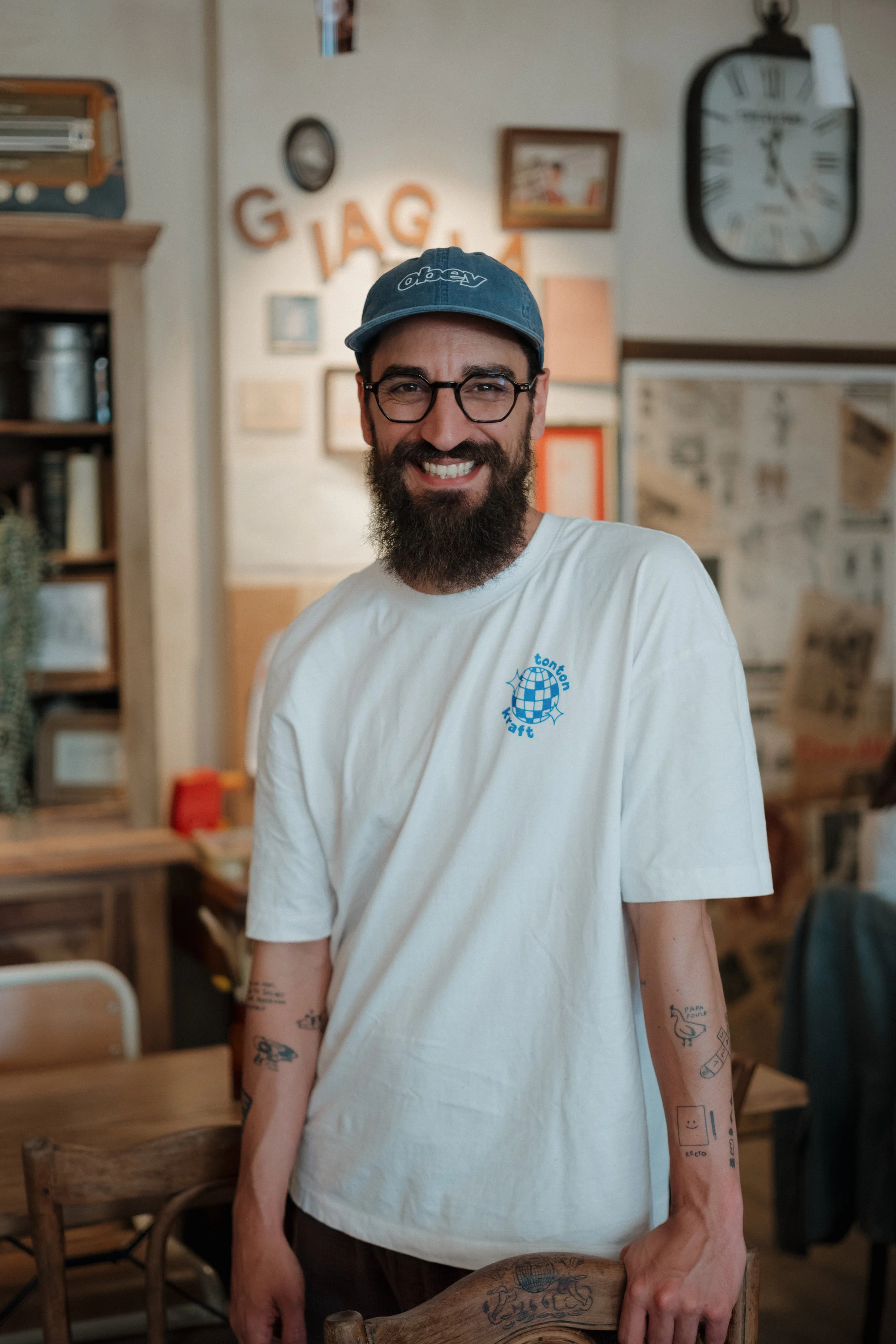 Un homme souriant portant une casquette et des lunettes, avec des tatouages sur bras, dans une pièce décorée avec des cadres et une horloge murale.