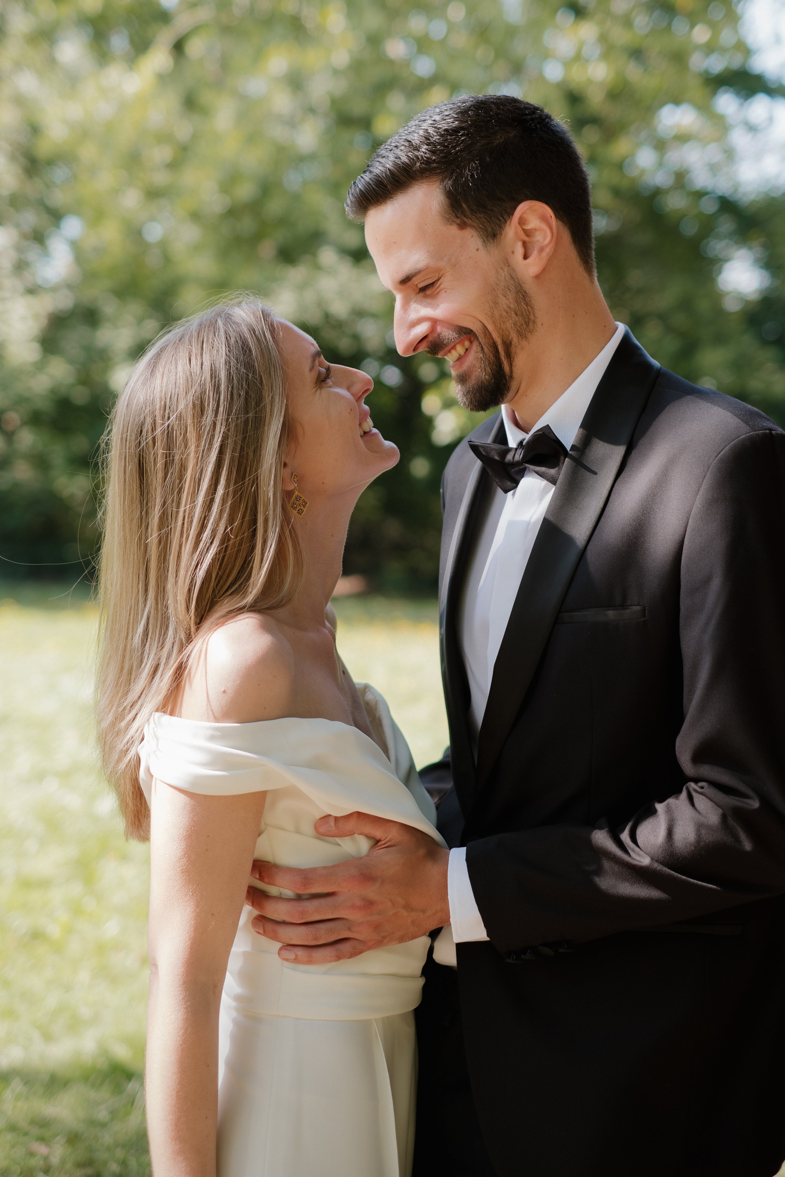 Un couple en vêtements de mariage, la femme en robe blanche et le homme en smoking, se regardant tendrement lors d'une séance photo en plein air, entourés d'arbres et de lumière naturelle.