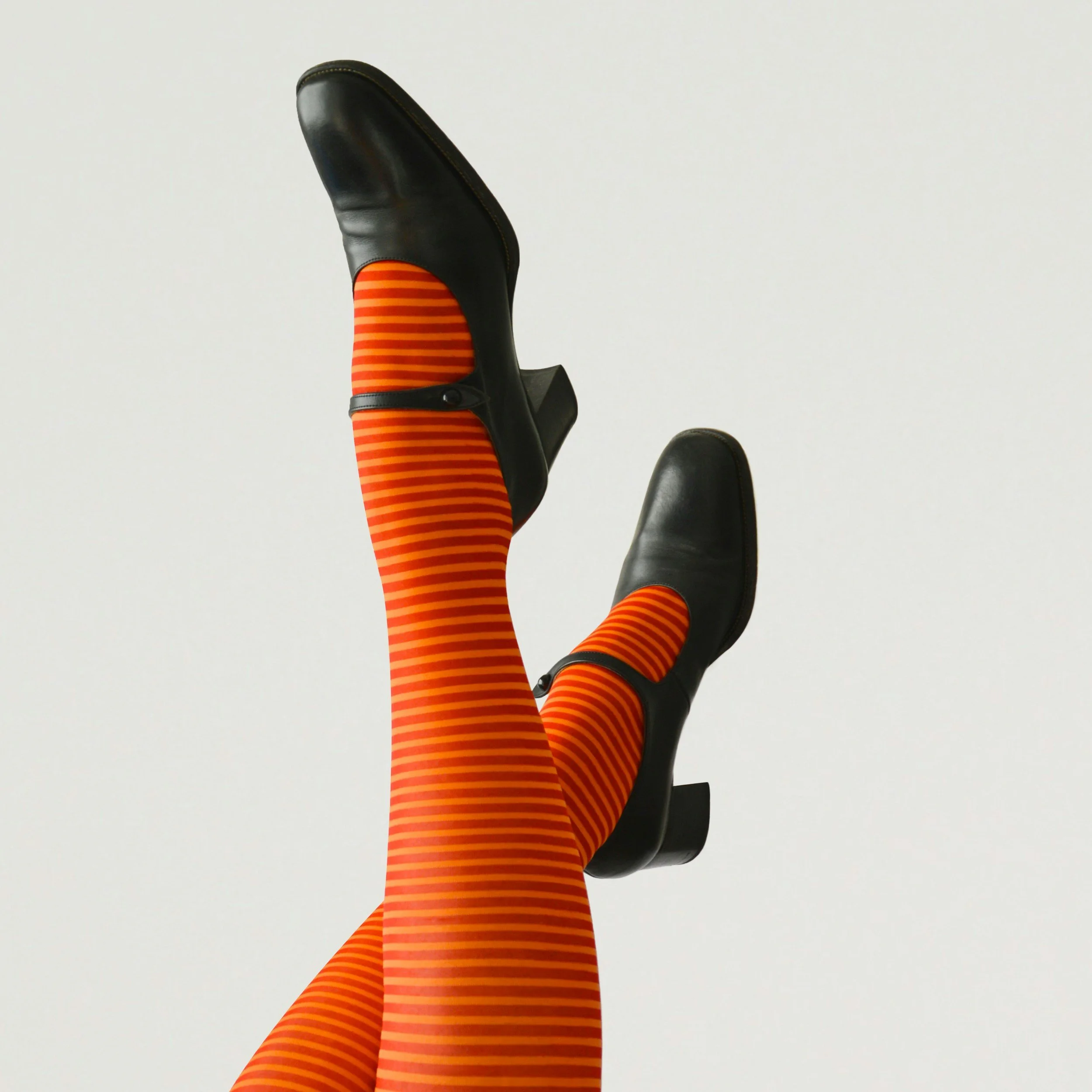 A person's legs wearing orange and black striped tights and black heeled shoes, with one leg raised against a plain white background.