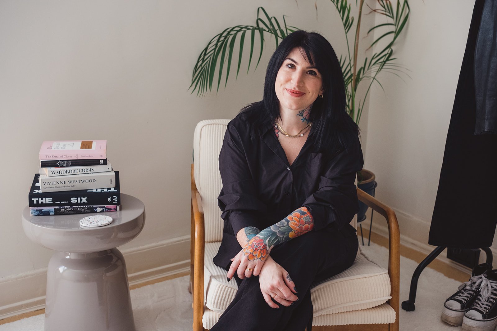Tattooed woman in black clothing sitting beside a stack of fashion books and a potted palm