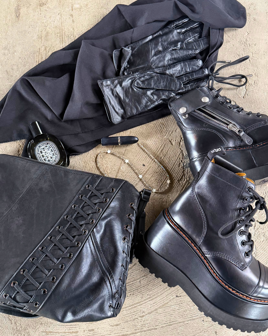 All-black fashion accessories arranged on a beige surface, including platform combat boots, leather gloves, black clothing, perfume, lipstick, necklace, and a handbag.