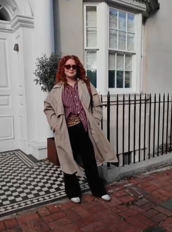Woman wearing sunglasses, a beige trench coat, striped shirt, black pants, and white shoes standing outside a townhouse.