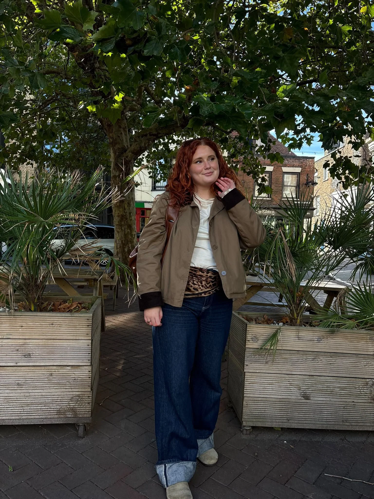 Red-haired woman posing outdoors between raised planters beneath a leafy tree, wearing a tan jacket and wide-leg jeans.