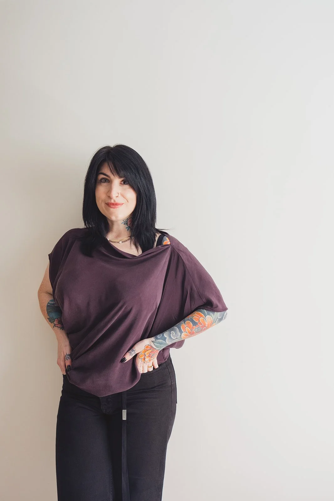 Woman with black hair and colorful floral tattoos wearing a plum top and black pants against a neutral wall