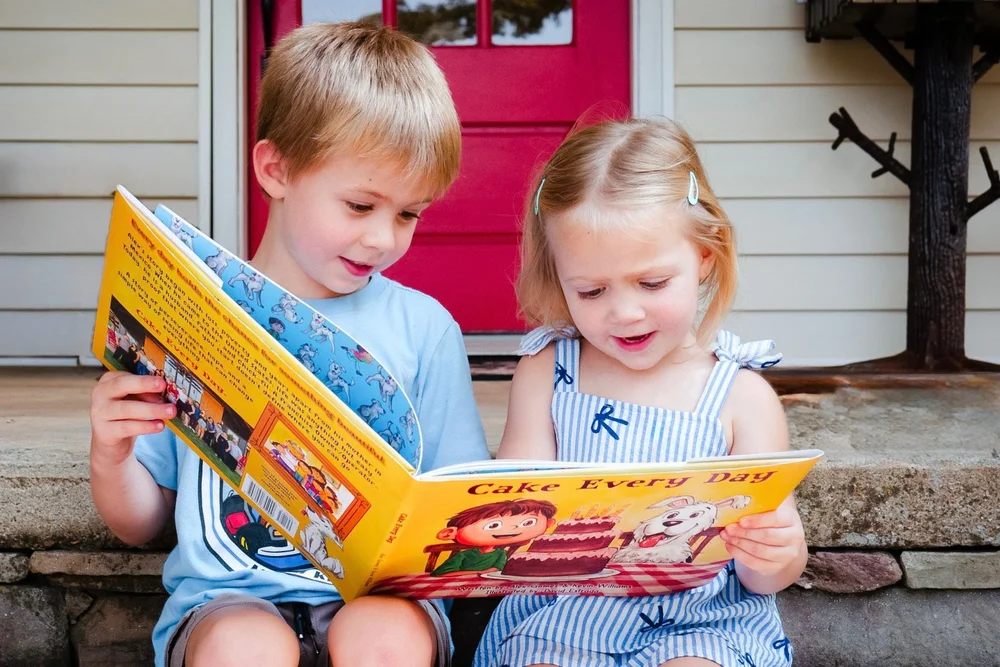 Two young children reading Cake Every Day together on a front porch