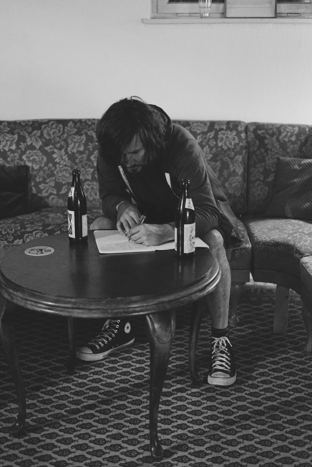 Person with dark hair, wearing a dark hoodie and Converse sneakers, sitting on a patterned sofa, leaning over a small wooden table, writing in a notebook with two beer bottles on the table.