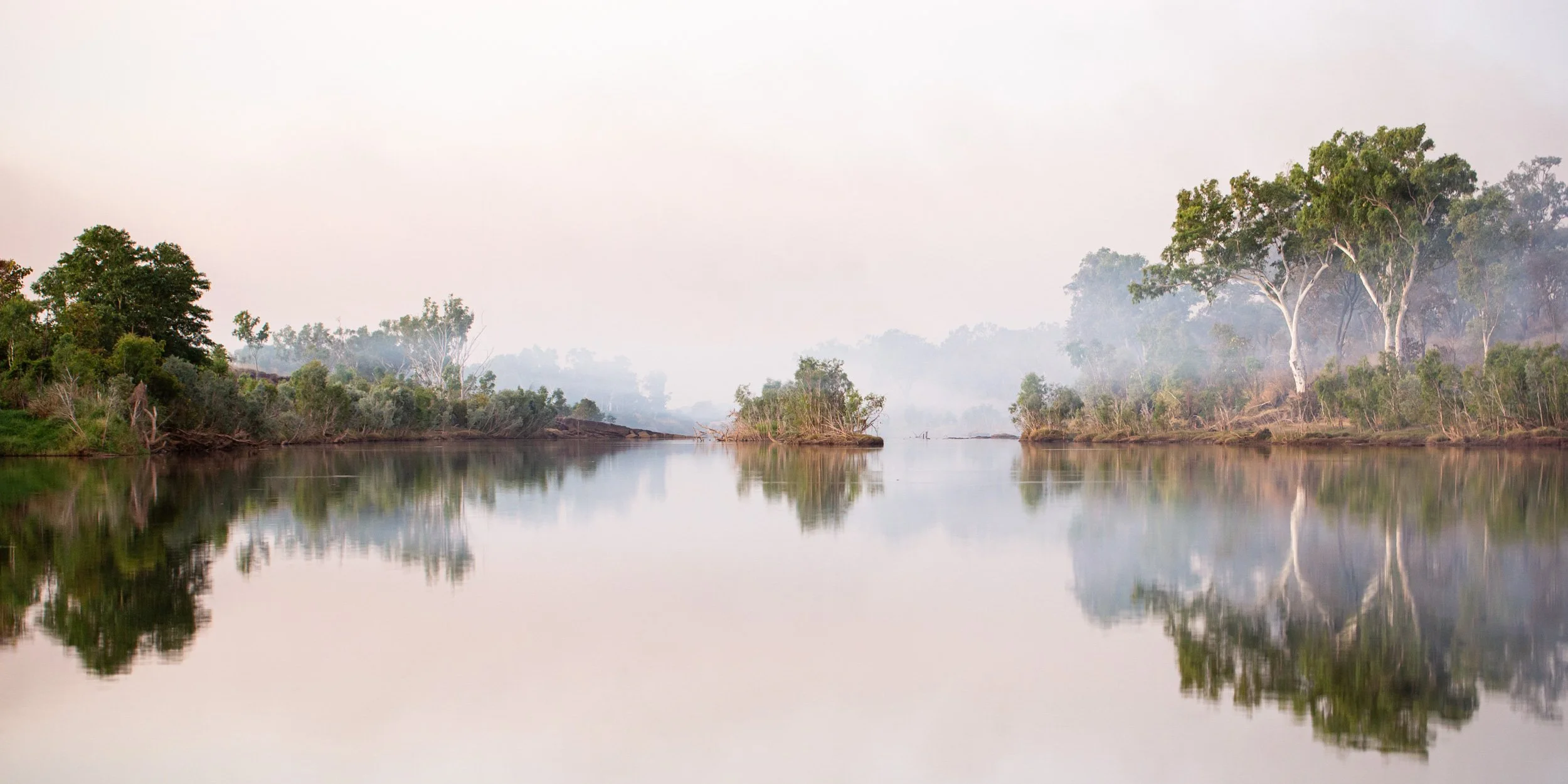 Smoke settles gently across the still waters of the lower Dunham River, softening the distant woodland into layers of muted light. 