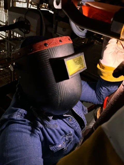 Person welding in a workshop, wearing a protective welding helmet, denim jacket, and gloves, with welding equipment in the background.