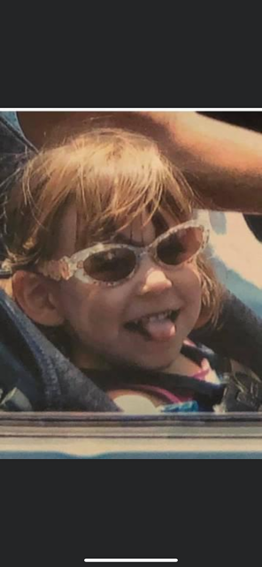 A young girl with light brown hair wearing pink sunglasses, sticking out her tongue and smiling while sitting in a stroller.