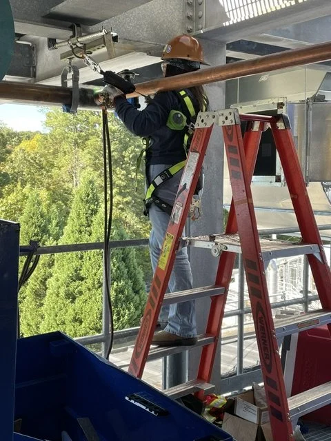 A construction worker wearing a safety helmet and harness stands on a ladder, working on overhead equipment at a construction site with trees visible in the background.