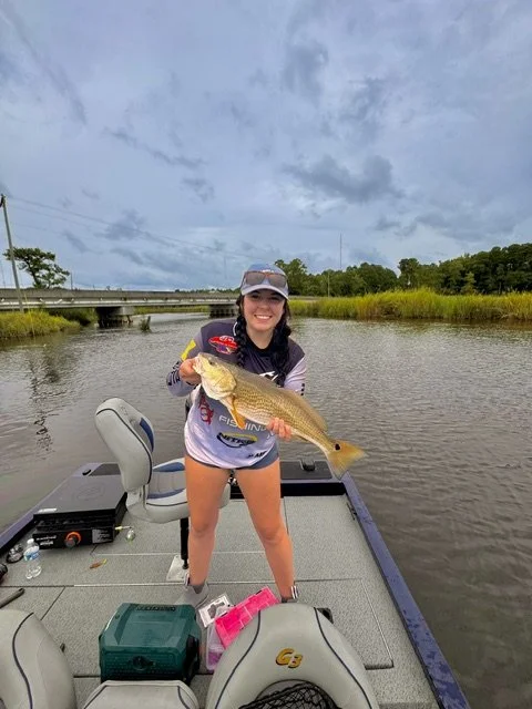 Young woman smiling on a boat holding a large fish, with a bridge and river in the background under cloudy sky.