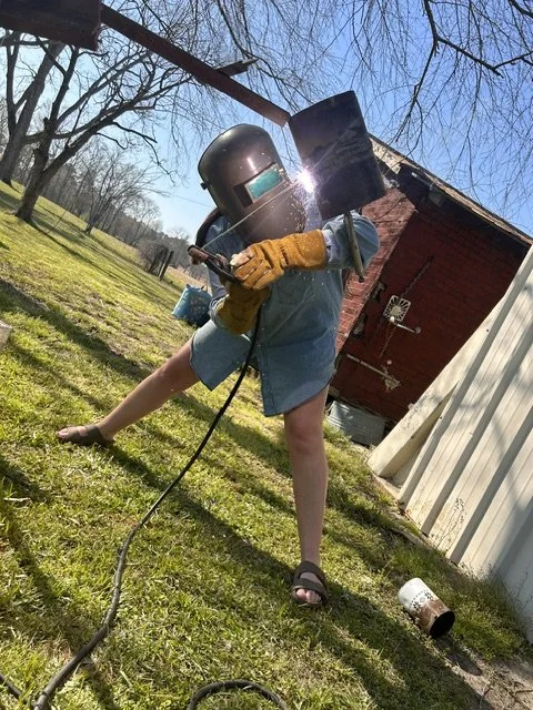Child in protective welding helmet and gloves welding outdoors in a grassy yard with a red shed in the background.