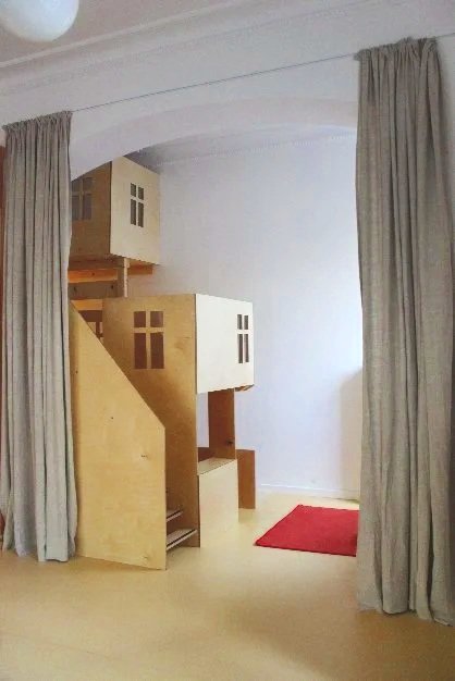 Wooden dollhouse with multiple rooms and windows, situated next to beige curtains in a room with a red rug on the floor.