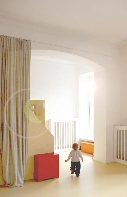 Child walking in a bright room with white walls, a window, beige curtains, and light-colored furniture.