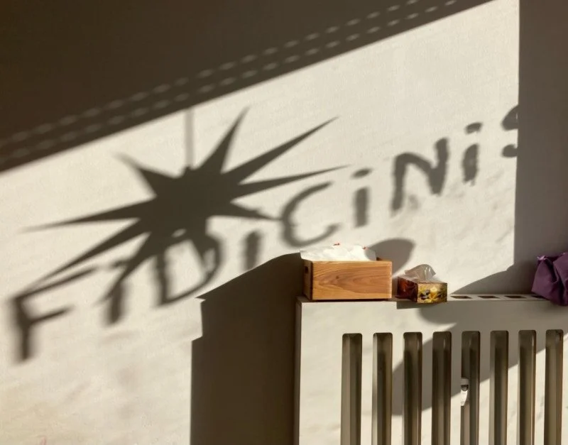 Sunlight casting shadows of a starburst shape and part of a word on a plain wall behind a beige radiator. A wooden box and small items sit on top of the radiator.