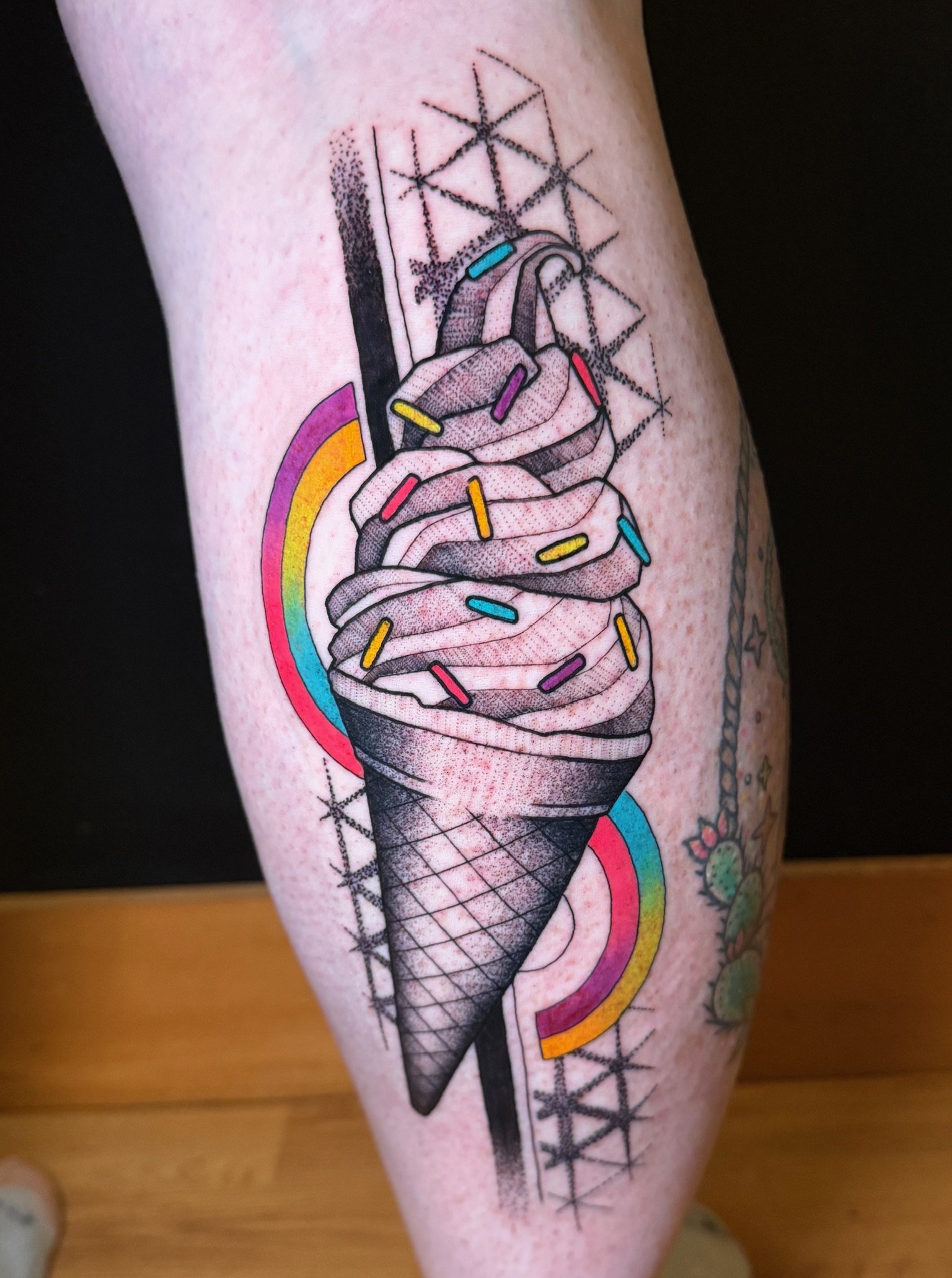 Tattoo of a stylized ice cream cone with colorful sprinkles and a rainbow, with geometric and abstract design elements.