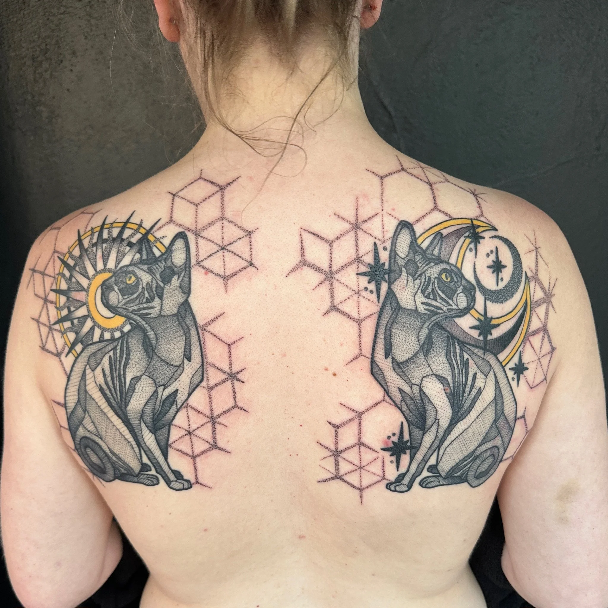 Back of a person's neck and shoulders with tattoos, including geometric patterns and animal imagery, against a dark background.