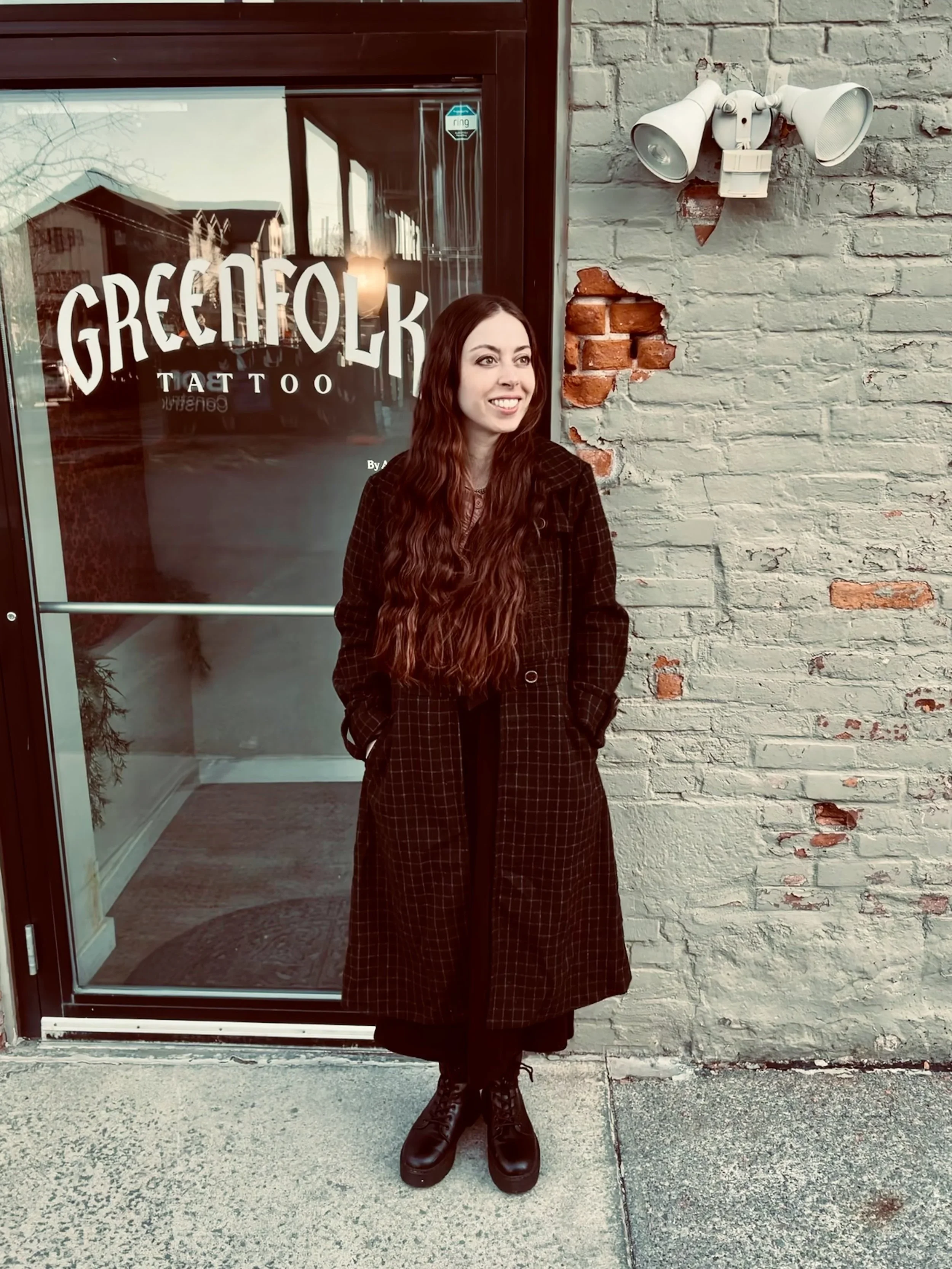 A woman with long, wavy red hair standing outside a brick wall building with a glass door that has the words 'GREEFOLK TATTOO' written on it. She is wearing a long, dark plaid coat, black dress, and black combat boots, with her hands in her coat pock