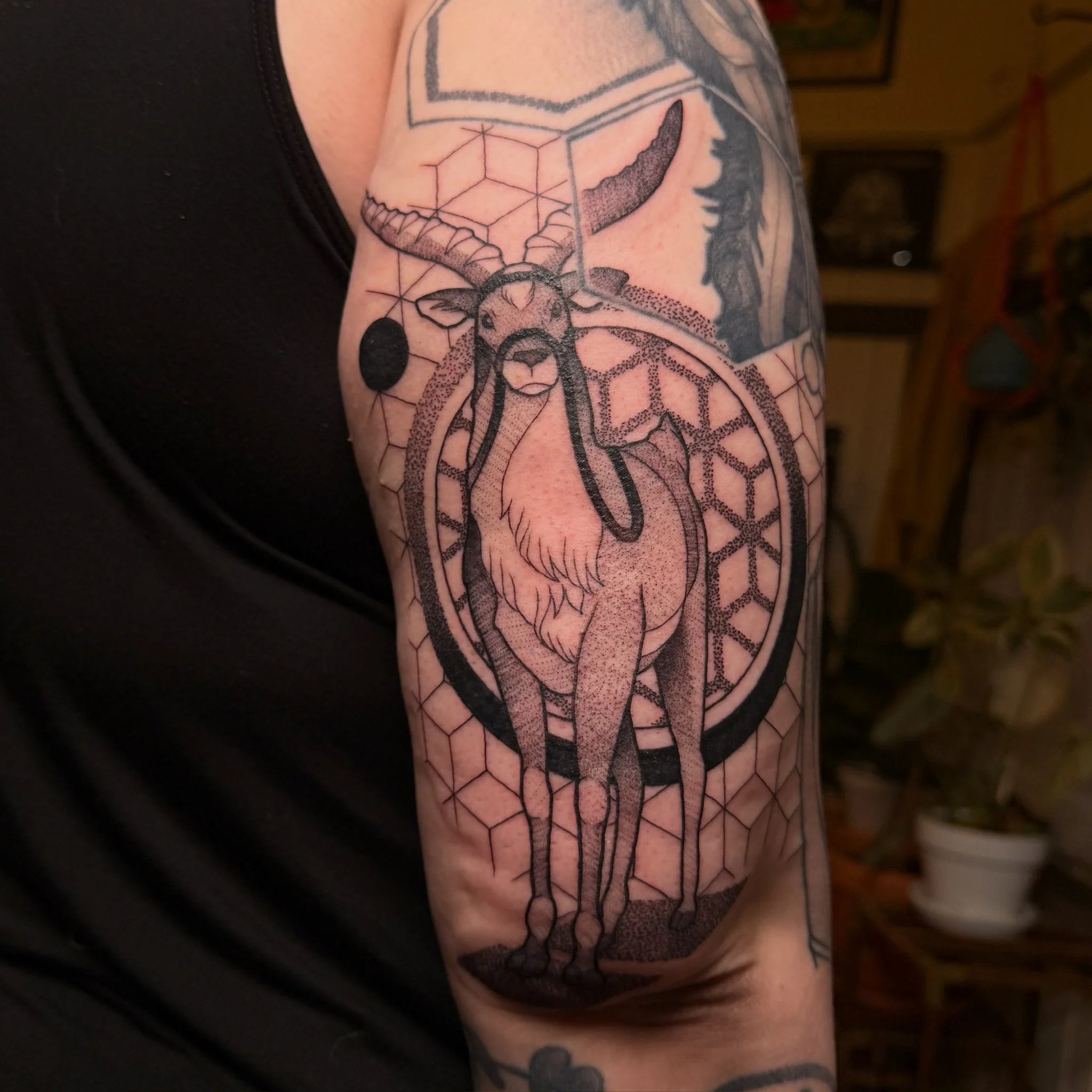 Close-up of a person's upper arm and shoulder displaying a geometric tattoo featuring a goat's head with horns and an abstract design incorporating cubes and hexagonal patterns.