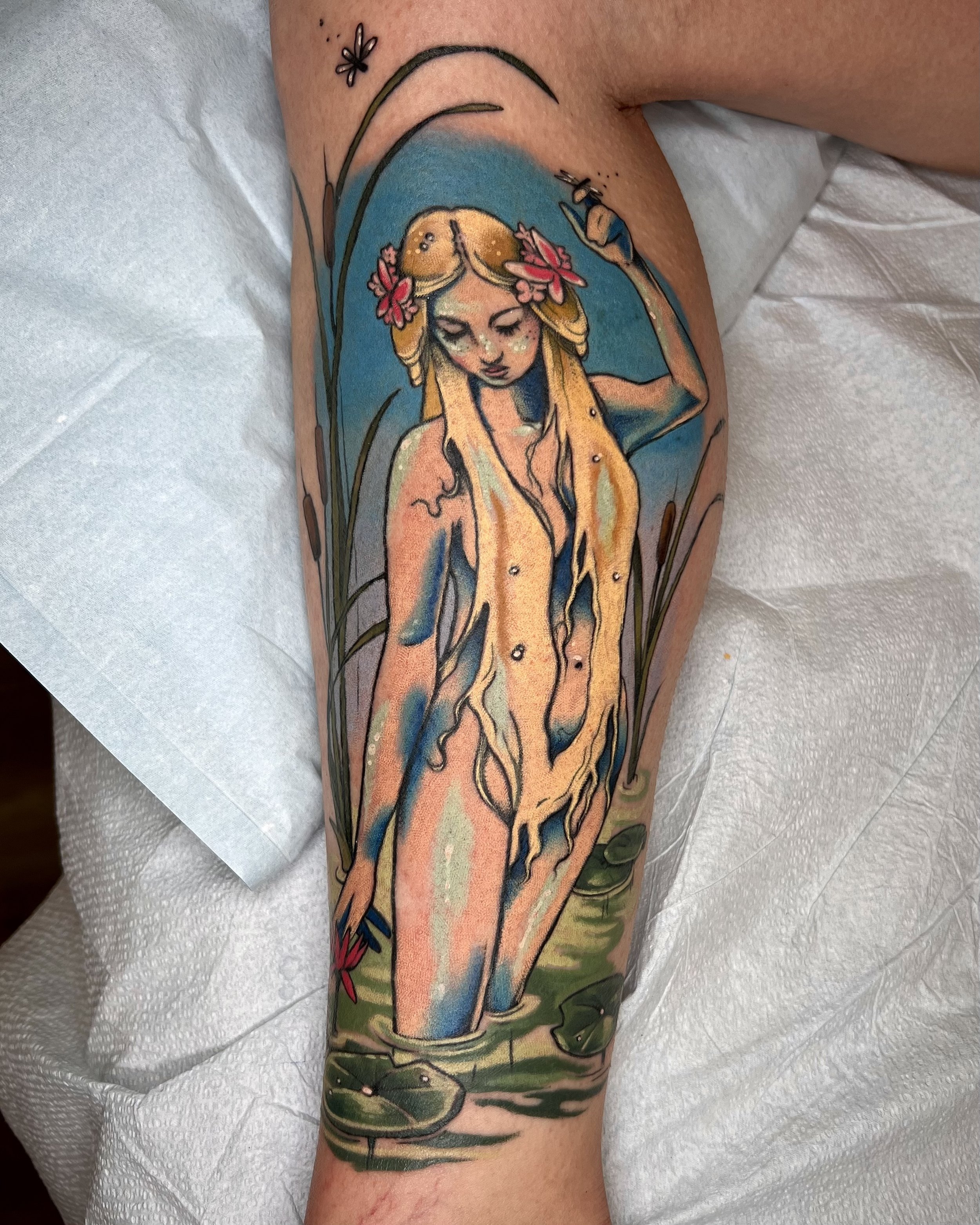Tattoo of a mermaid with long blonde hair, pink flowers in her hair, holding a fishing pole, surrounded by water and lily pads.
