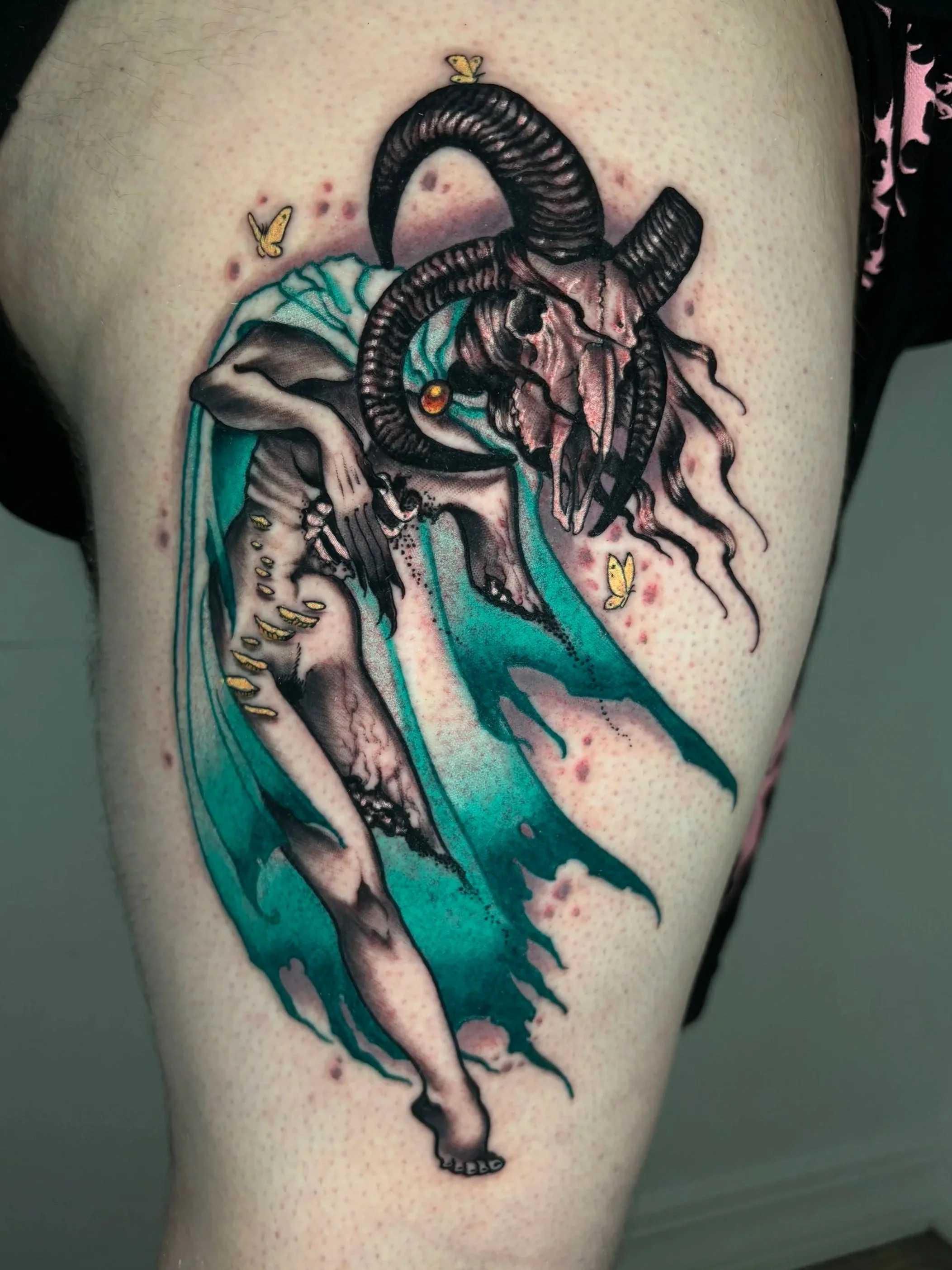 Colorful tattoo of a ram's head with horns, fangs, and a skull on the person's arm, with small yellow butterflies around it.