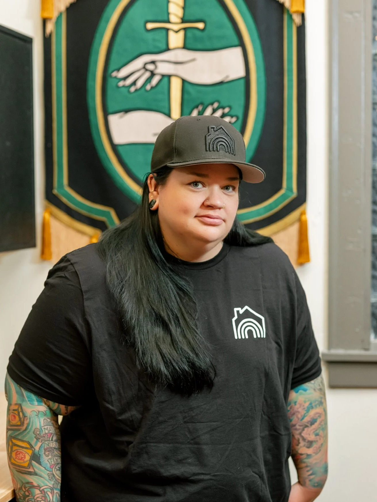 A woman with black hair and tattoos on her arms, wearing a black shirt and a gray cap with a logo, standing in front of a black banner with a green and white emblem.
