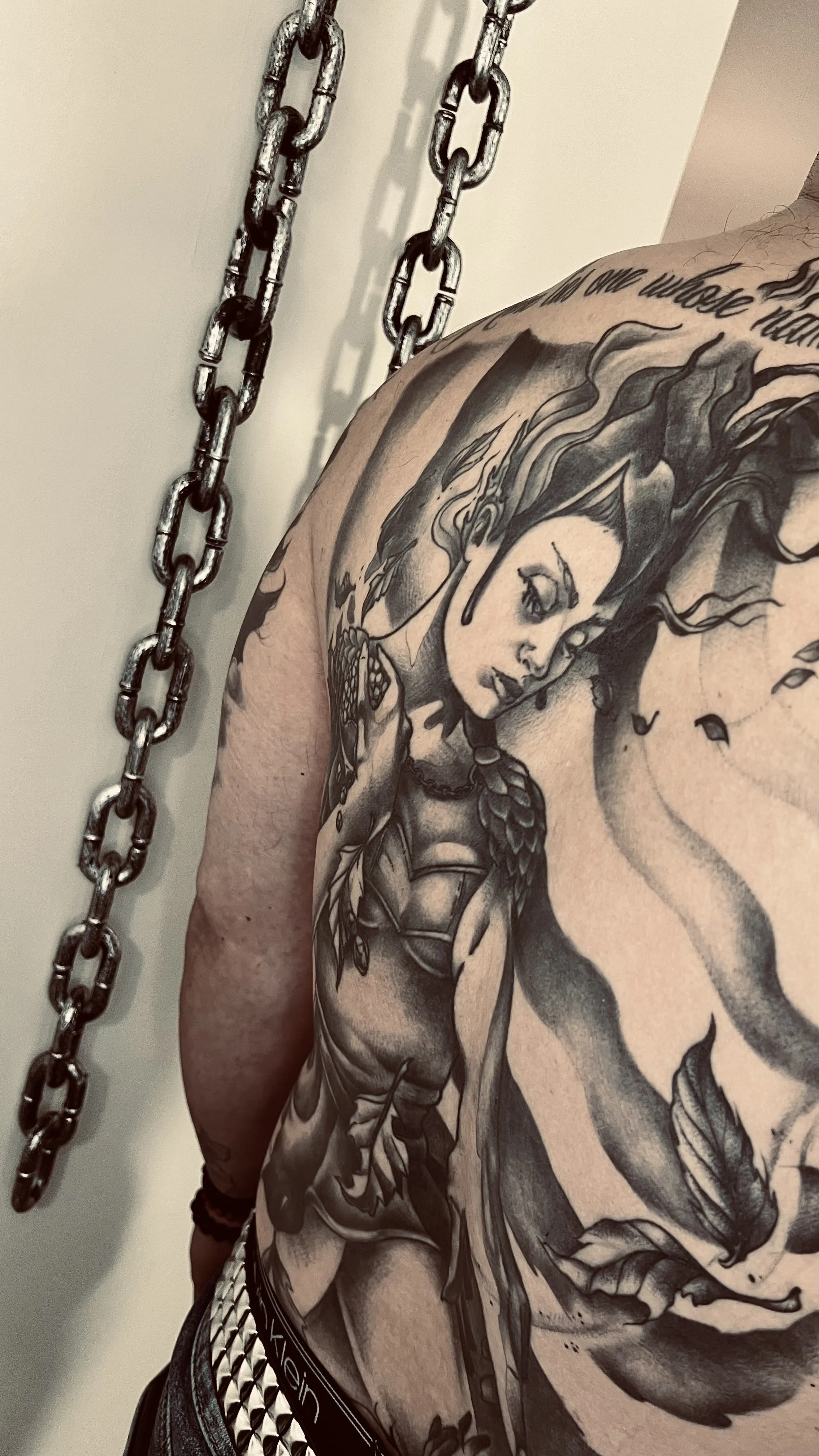 A person's shoulder and upper arm with a large black and gray tattoo of a woman with flowing hair, earrings, and detailed facial features. There is a chain hanging in the background and part of a bracelet is visible on the wrist.