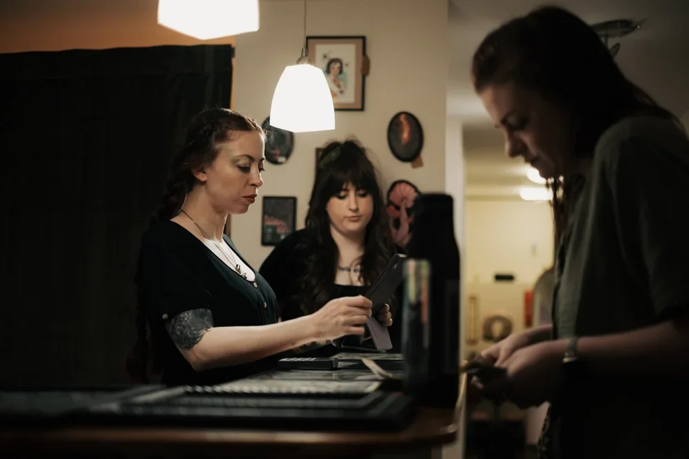 Three women standing at a kitchen counter, looking at their phones, with warm lighting and framed pictures on the wall behind them.