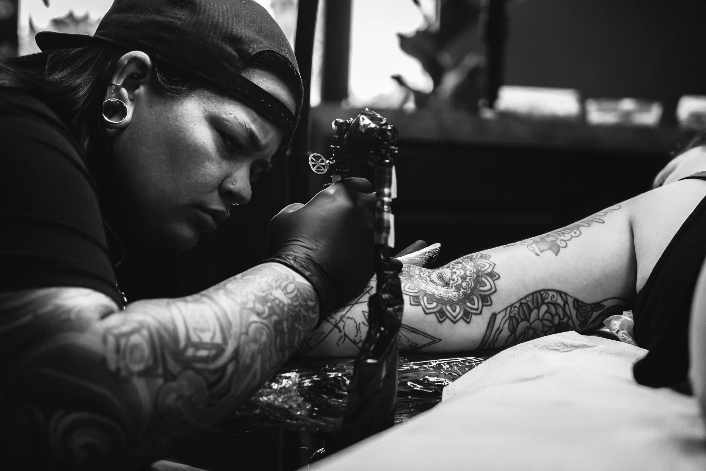 A tattoo artist with piercings and a cap is applying a tattoo to a person's arm.