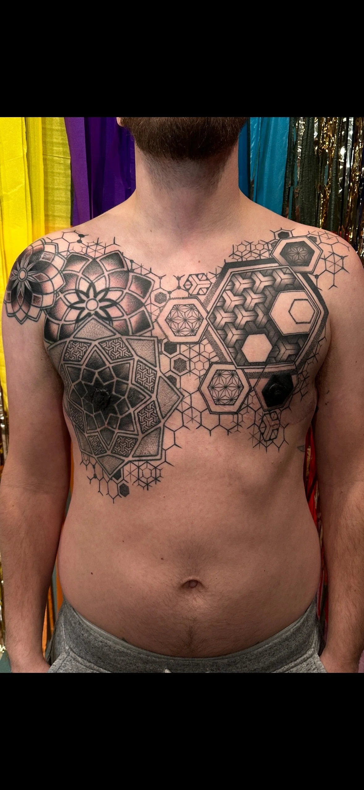 A man with a beard shows a geometric tattoo on his chest and shoulders, with hexagon and floral patterns, in front of a colorful backdrop.