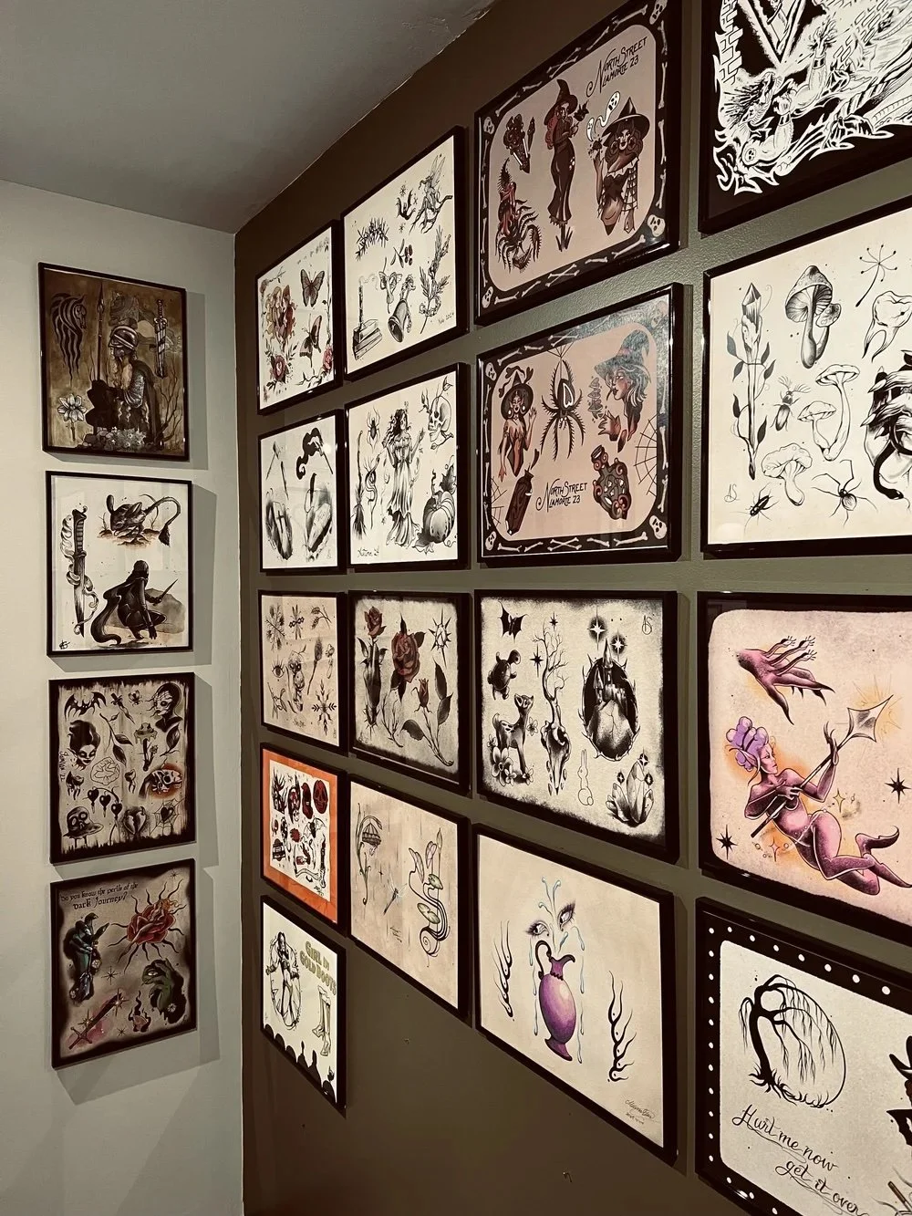 Collection of framed tattoo flash art prints on walls, featuring traditional and neo-traditional designs including mythological figures, animals, skulls, and whimsical characters in black, white, and muted colors.
