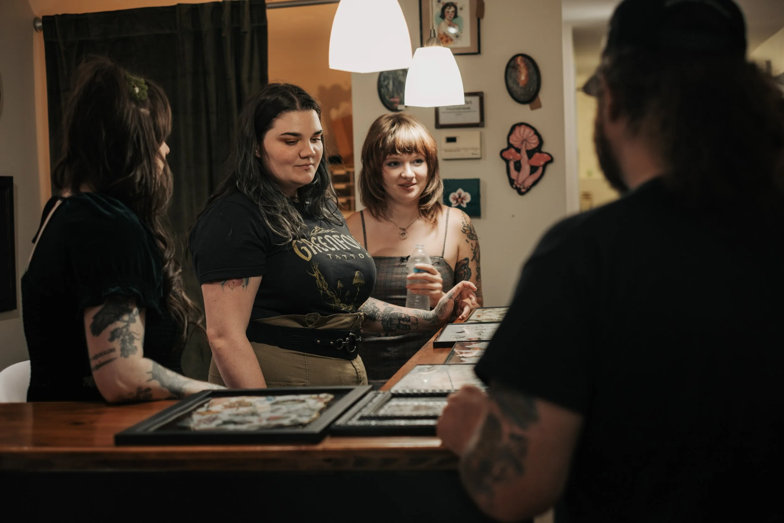 Four people at a bar or restaurant looking at a menu or tabletop, with artwork on the wall in the background, including a framed picture and decorative plates.