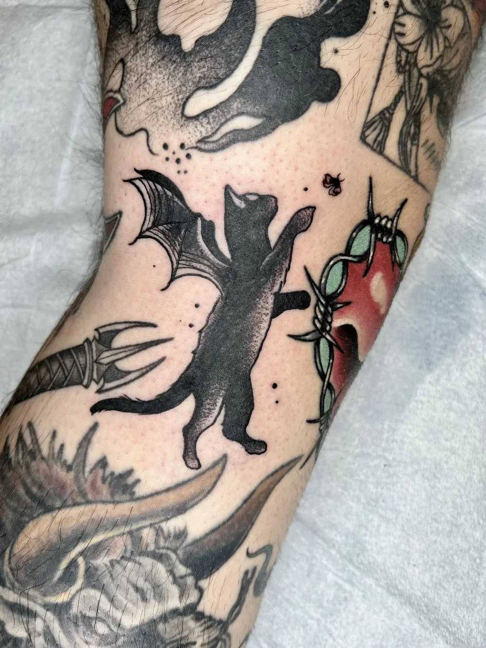 Tattoo of a black cat with bat-like wings reaching for a small bee, with a red heart with green leaves nearby, on a person's arm.