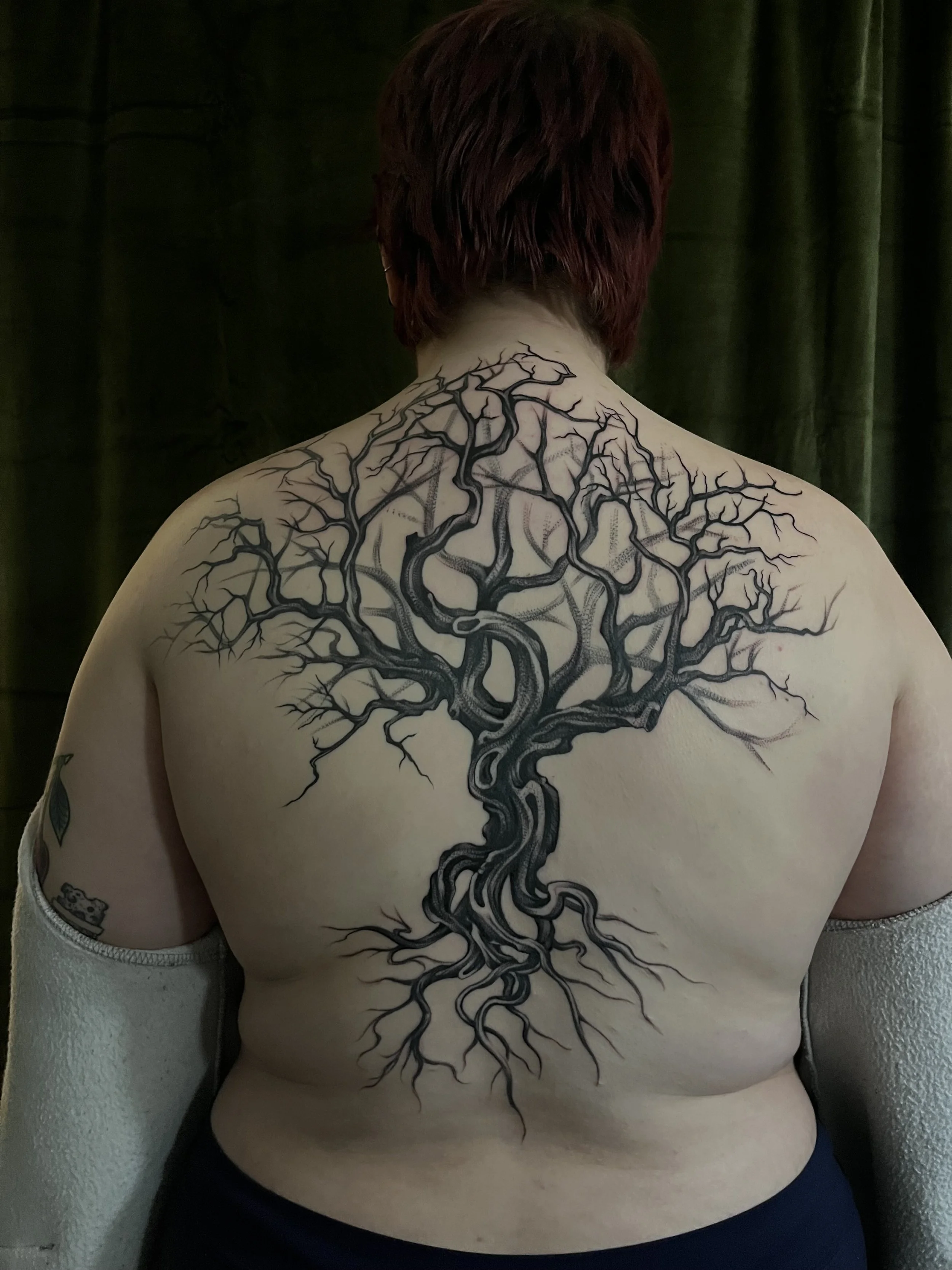 A person with short, dark hair showing their back, featuring a large black tattoo of a tree with twisting and branching limbs covering their upper back and shoulders.