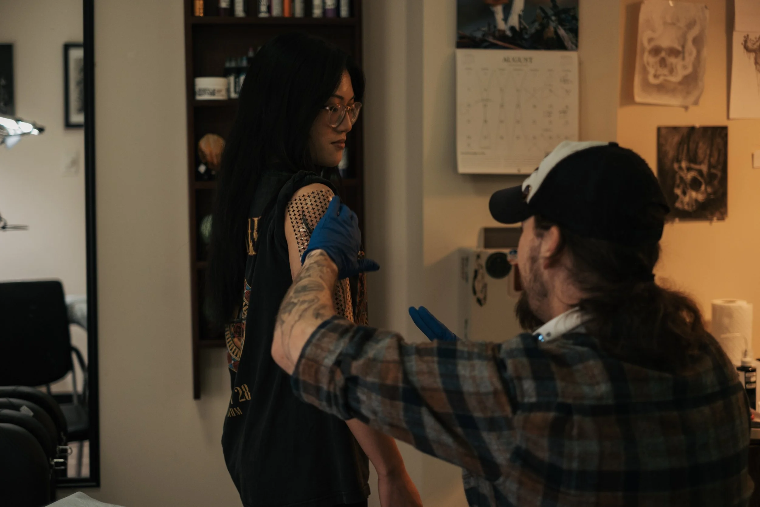 A woman with long dark hair and glasses sitting in a tattoo studio as a tattoo artist tattoos her arm.