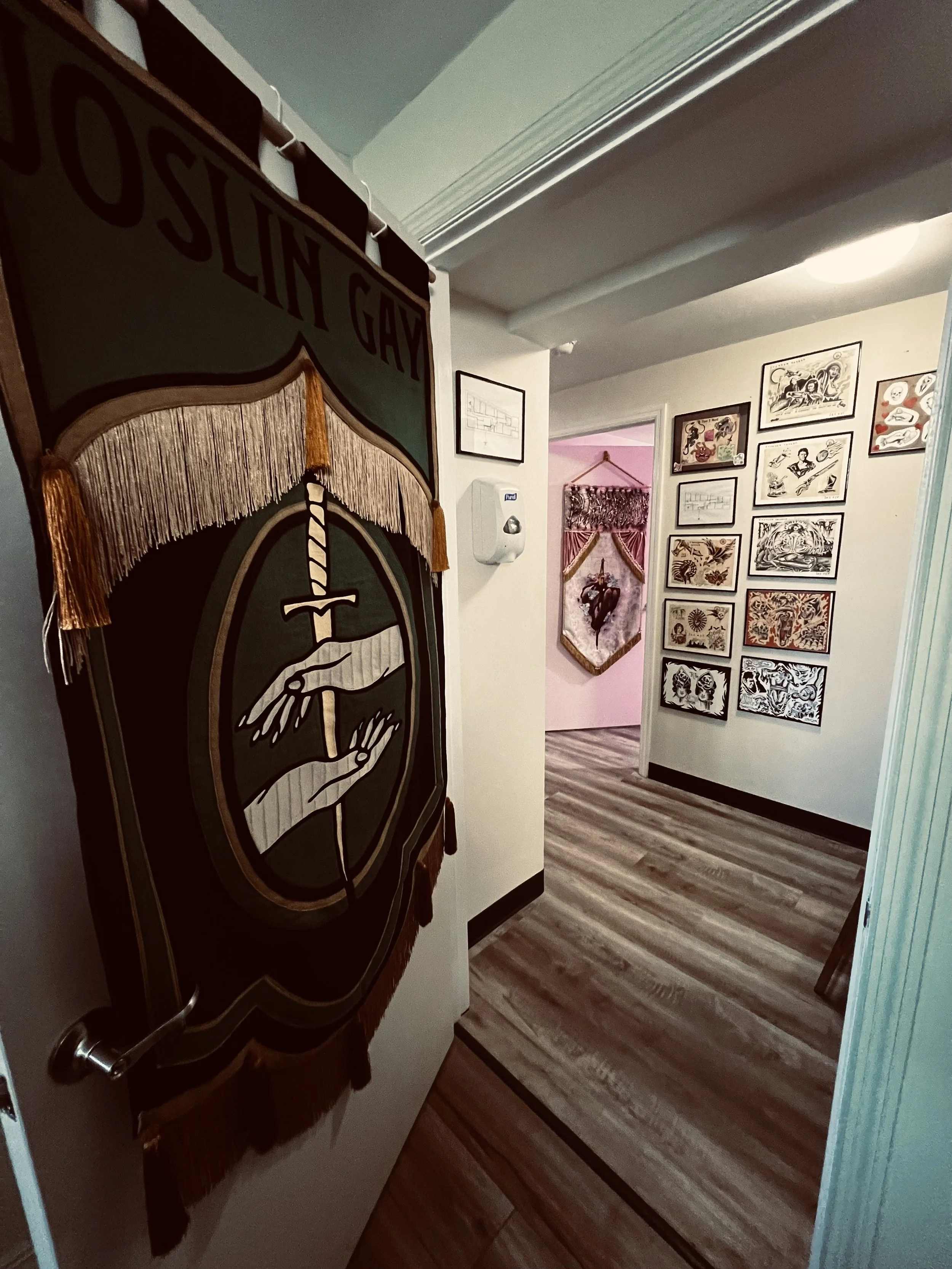 Hallway with a framed gallery wall of various artwork and photos, a pink wall with a hanging tapestry, and a wooden floor.