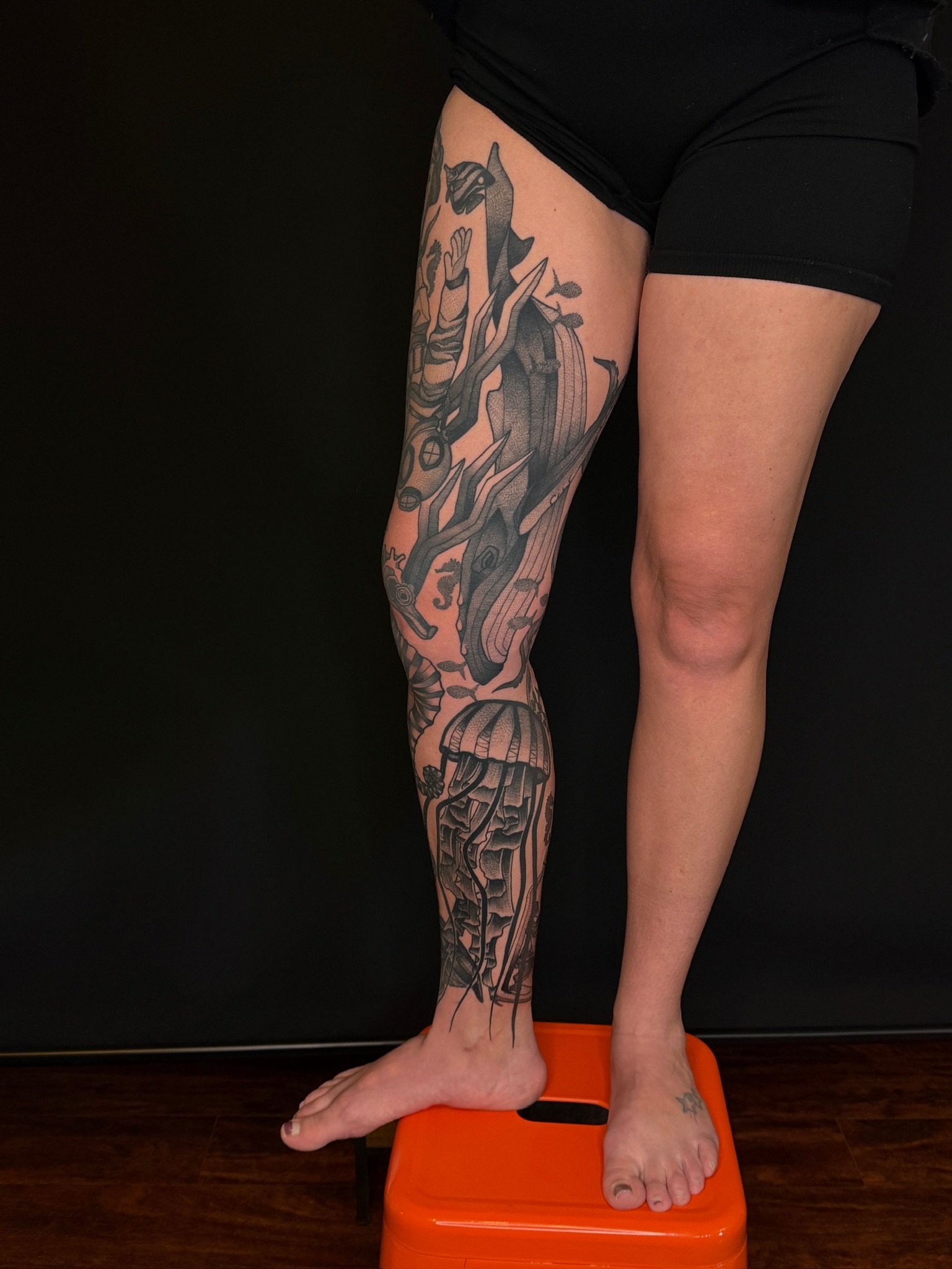 A person standing on an orange balance scale with one foot, with the other leg extended and resting on a black background. The person has a tattooed leg.