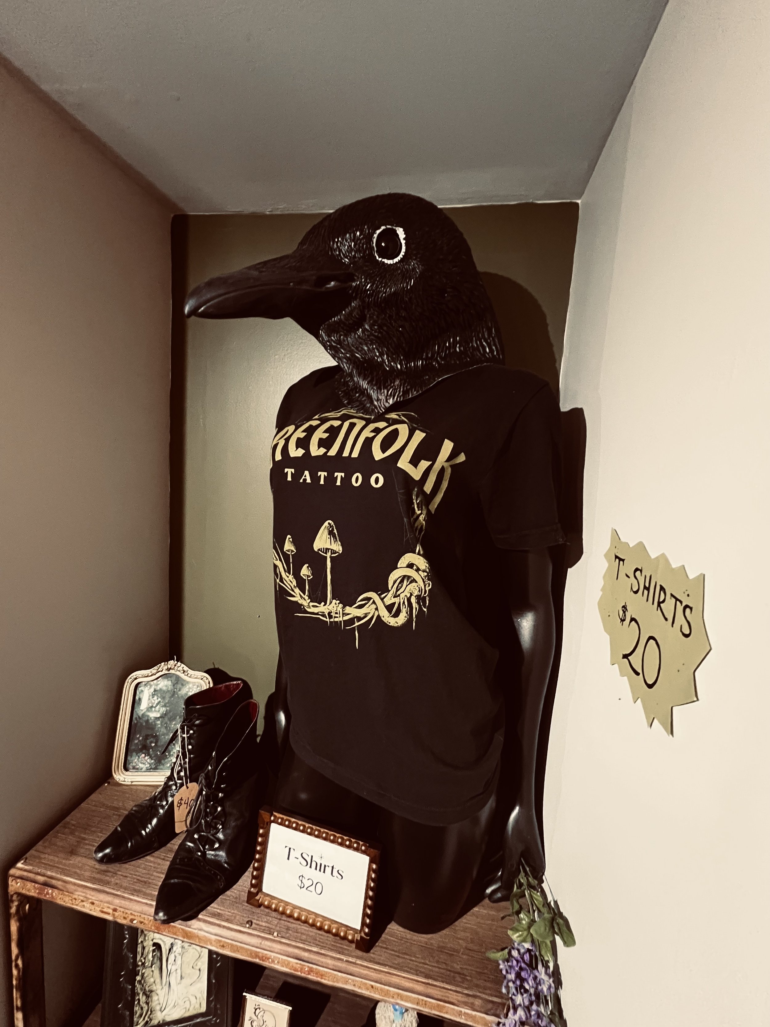 Mannequin with a bird mask wearing a black t-shirt that says 'GREENFOLK TATTOO' and stand with black leather boots, on table with a sign that says 'T-Shirts $20', in a retail display area.