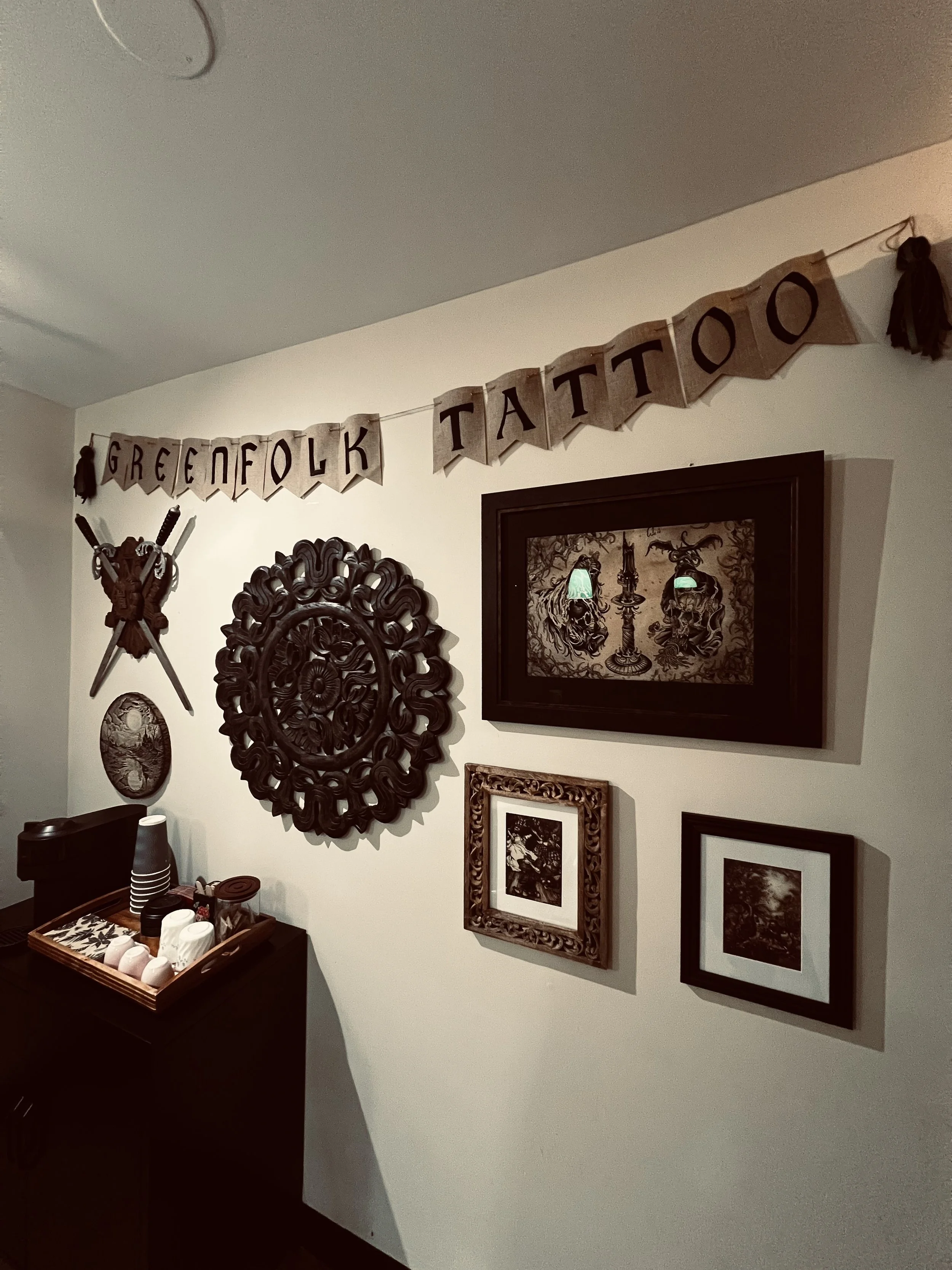 Decorative wall with framed pictures and a banner reading 'Greensfolk Tattoo' in a room with a table of cups and a decorative wooden coat rack.
