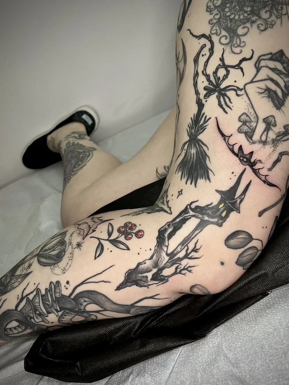 Close-up of a person's heavily tattooed arm and side torso, showing various black ink designs including a tree, a bat, berries, mushrooms, and abstract elements.