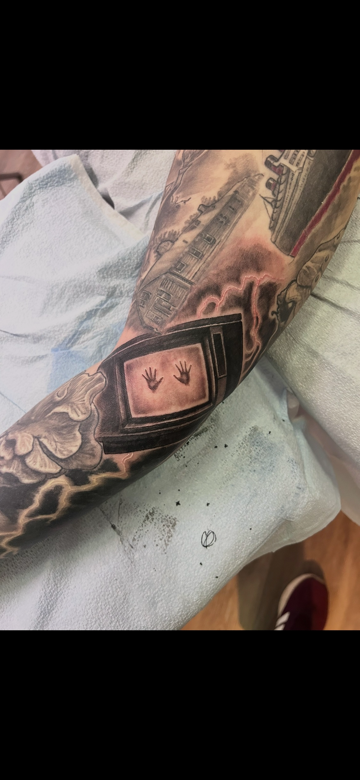 Tattoo on arm featuring a vintage television with two hands reaching out of the screens, surrounded by clouds and buildings in the background.