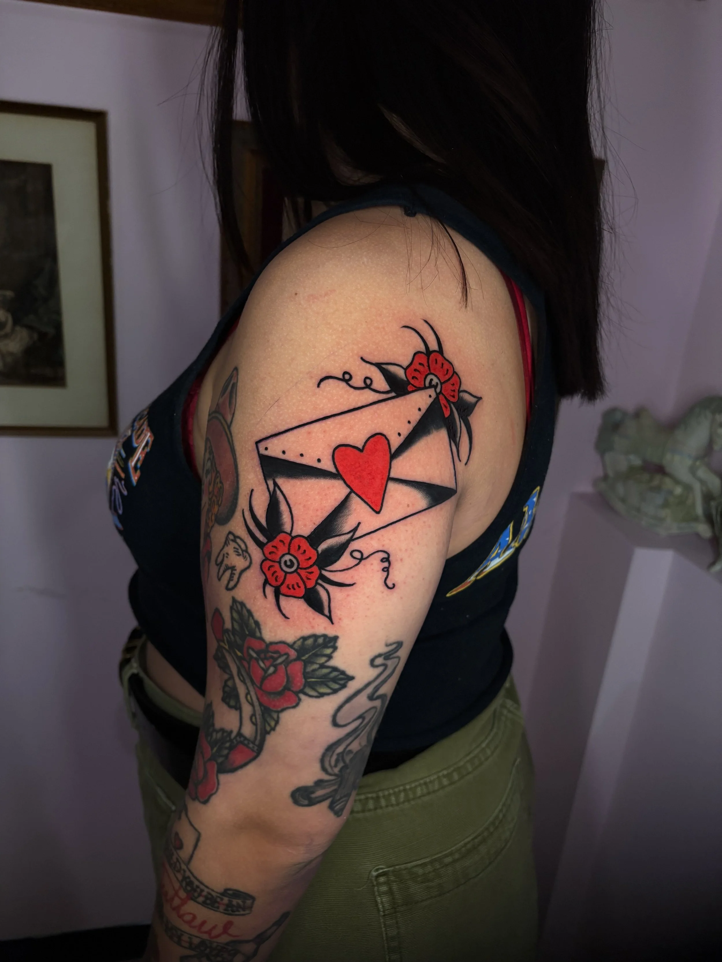 Close-up of a person's upper arm and shoulder with colorful tattoos, including a red flower, a red heart inside an envelope, and various other designs. The person is wearing a sleeveless shirt and olive green pants.