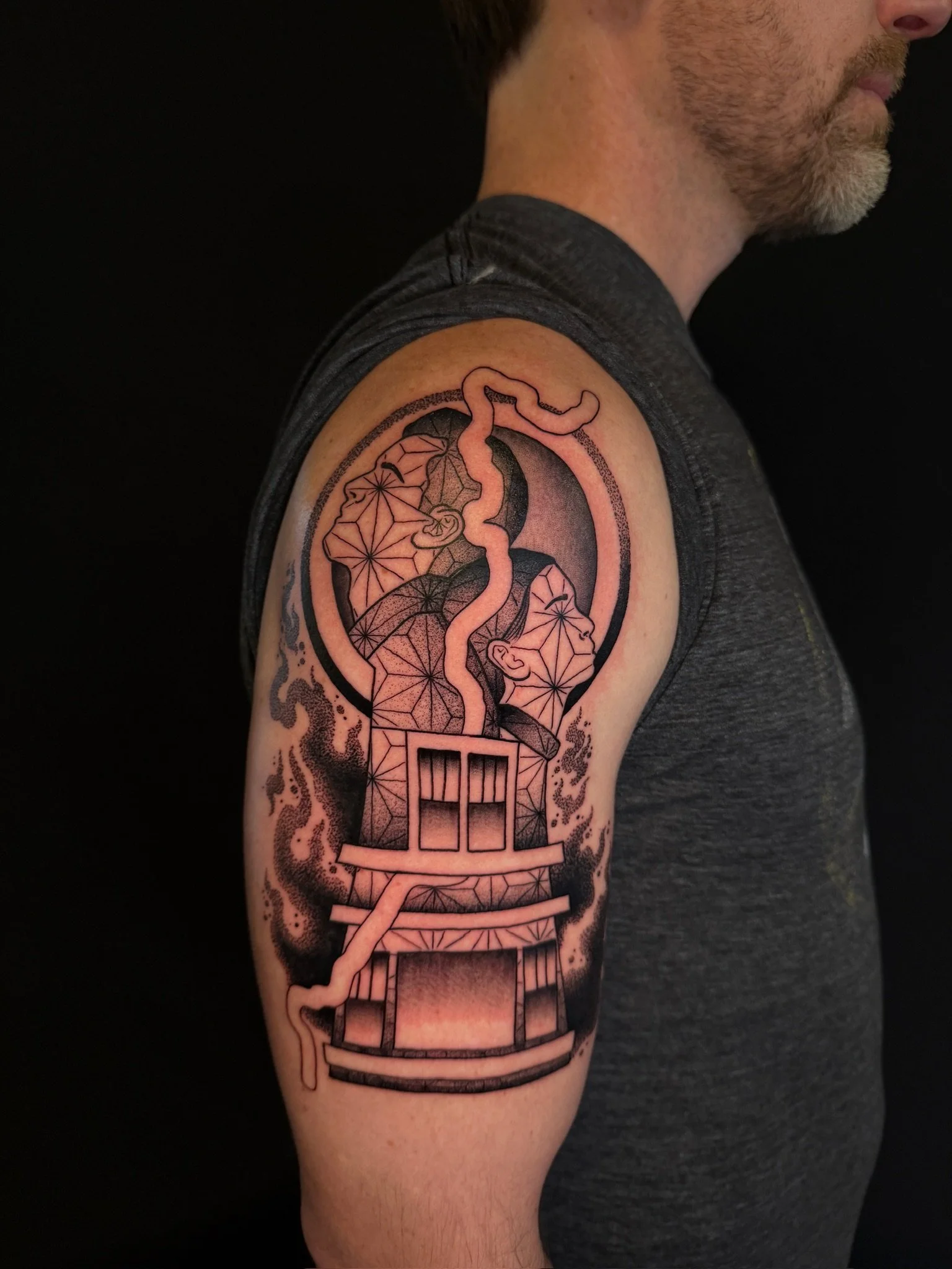 A tattoo on a man's upper arm featuring a circular design with stylized human faces and a snake intertwined with geometric patterns and a ladder. The tattoo uses black ink and detailed line work.