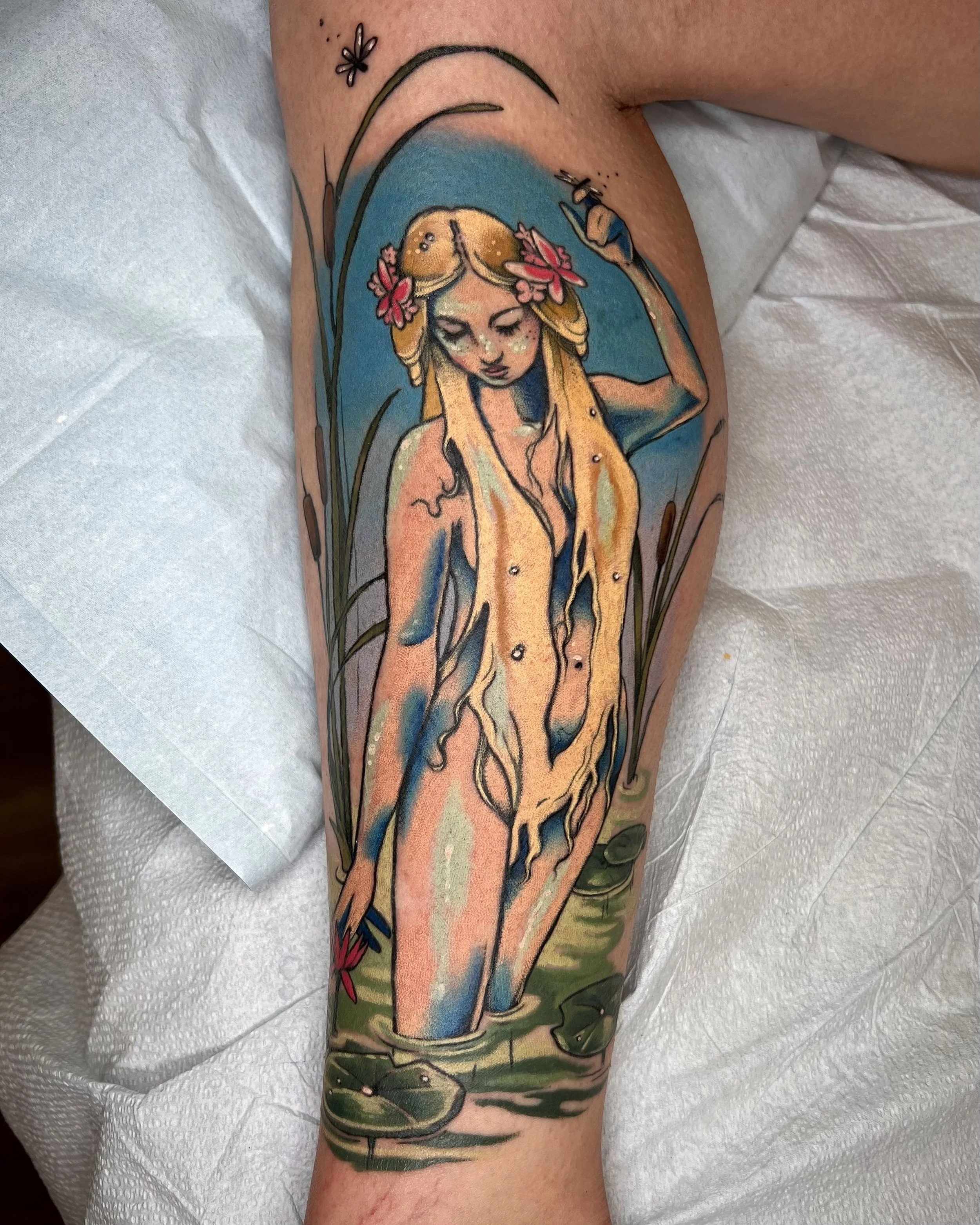 A colorful tattoo on a person's arm depicts a mermaid with closed eyes and long flowing hair, surrounded by grass, lilies, and a dragonfly.