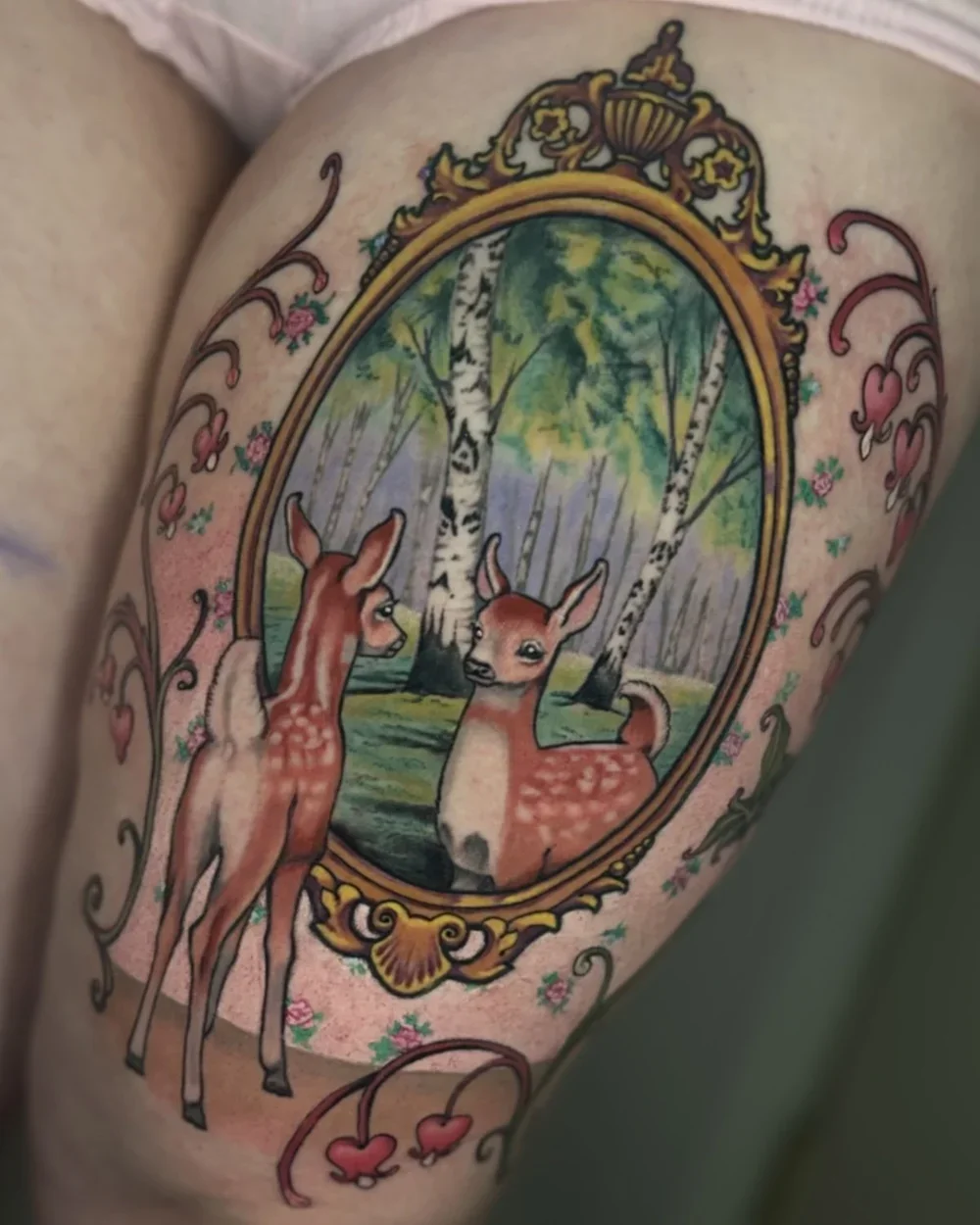 Tattoo of a framed painting of two deer in a forest, with heart and floral designs surrounding the frame.