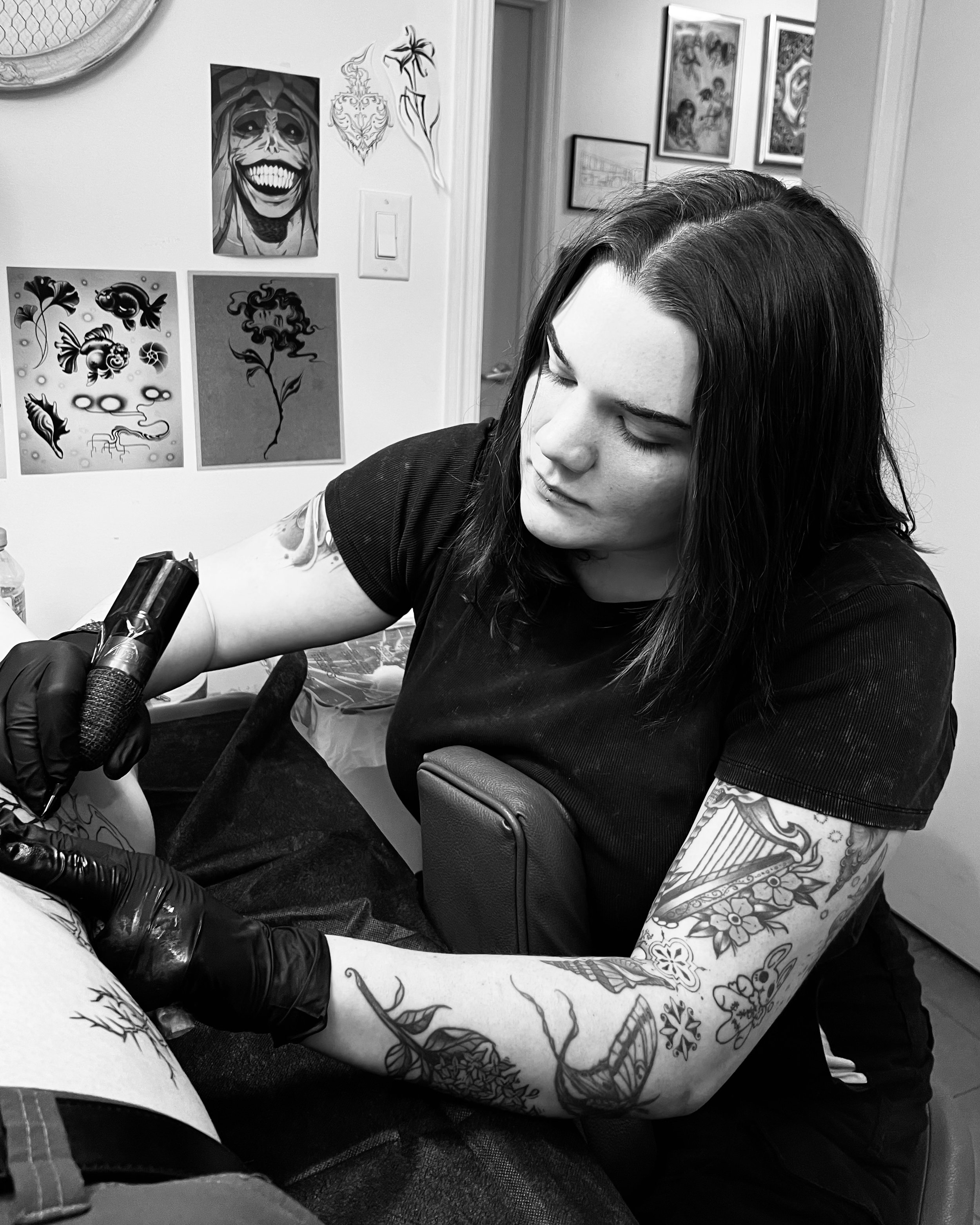 A woman with dark hair and visible tattoos on her arm is tattooing a design on someone's leg in an art studio. The wall behind features various artwork, including a smiling face and tattoo sketches.