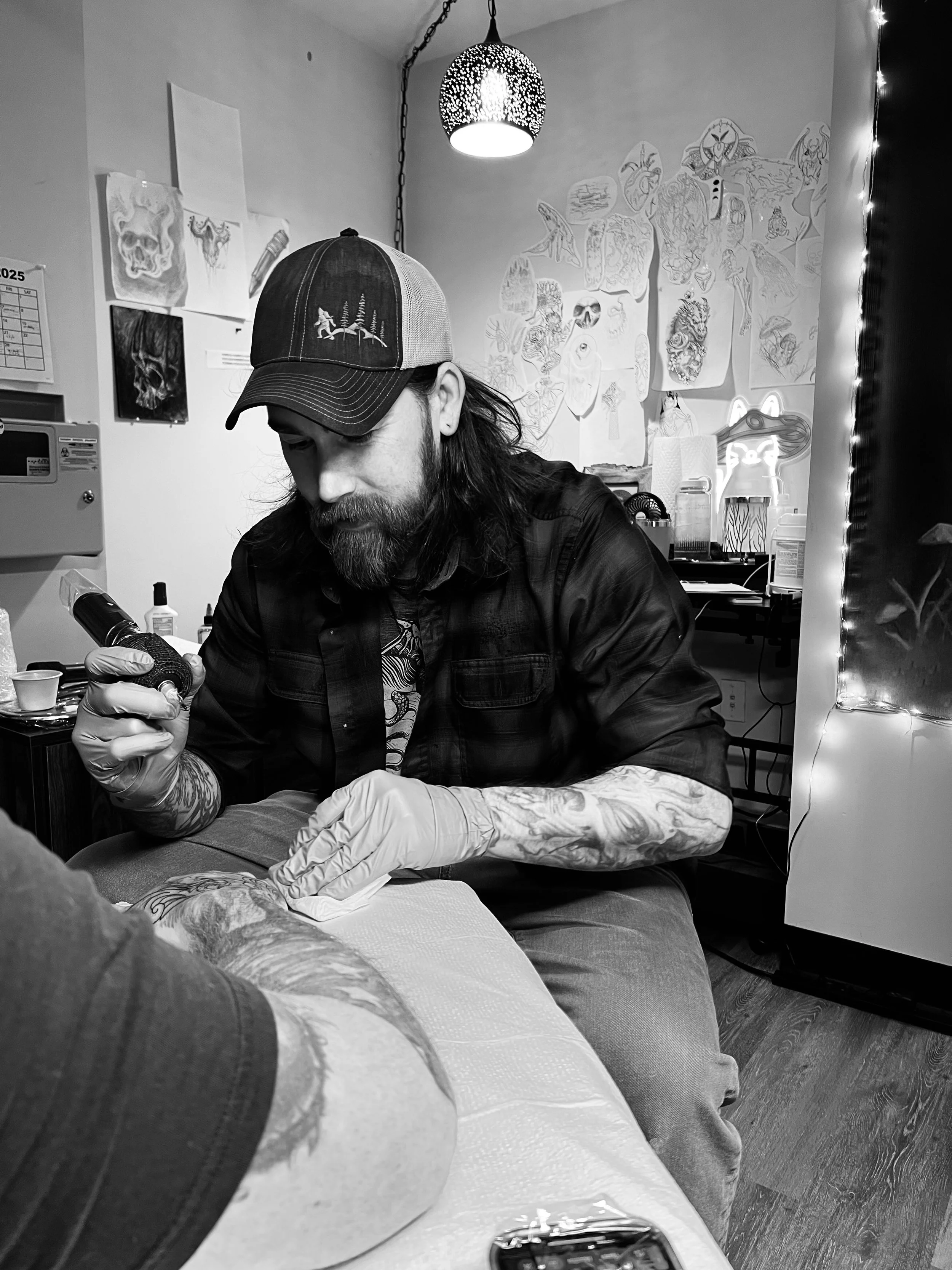 A tattoo artist with tattoos on his arms, wearing a cap with trees, working on a person's arm in a tattoo studio. The wall behind him has drawings and sketches, and there's a lamp hanging from the ceiling.
